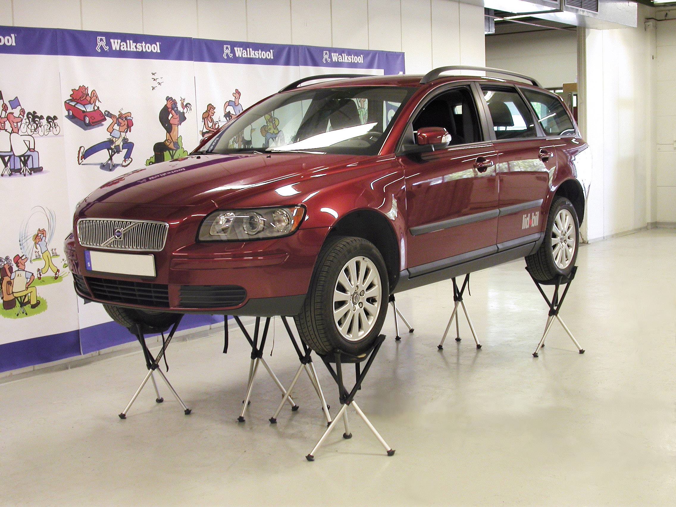 Une voiture rouge est posée sur plusieurs tabourets pliés dans une salle d'exposition. L'arrière-plan montre des dessins de bande dessinée sur le mur.