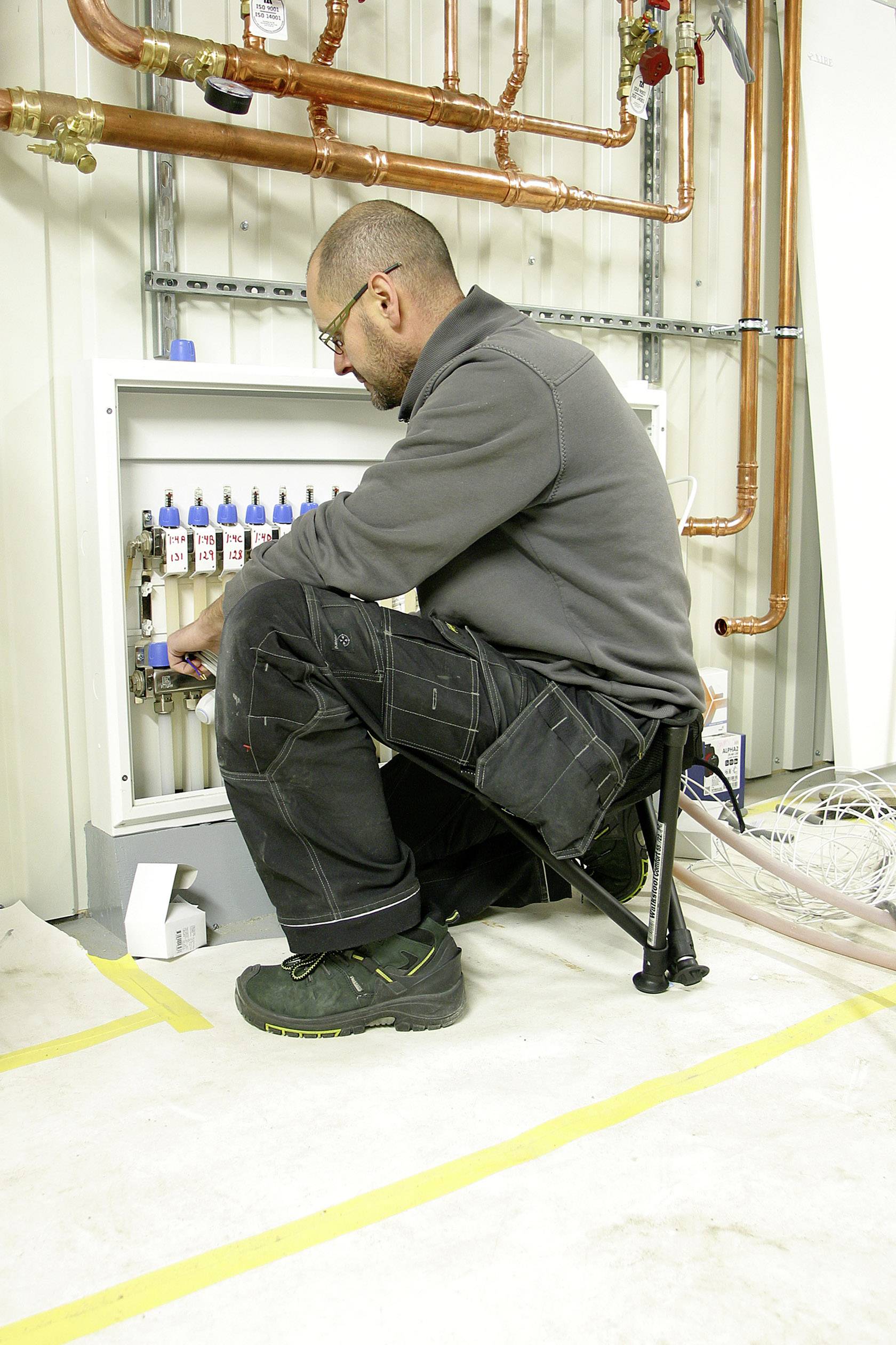 Un homme en combinaison de travail est assis sur un tabouret et travaille sur un coffret de distribution avec plusieurs tubes dans une salle technique.