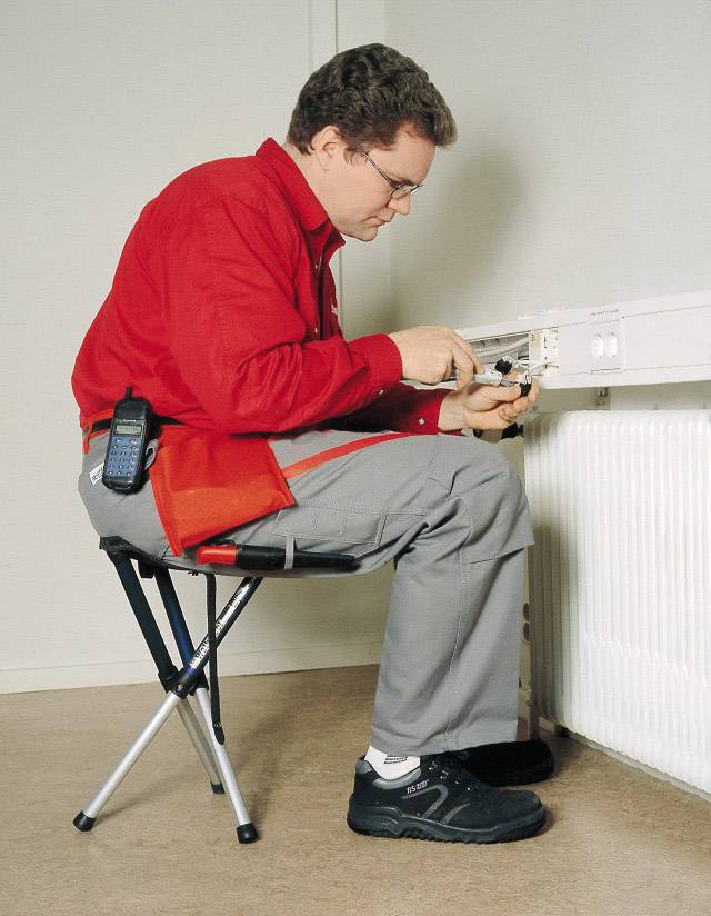 Un technicien en chemise rouge et pantalon gris répare une prise électrique tout en étant assis sur un tabouret pliant. Une radio portative est accrochée à son côté.