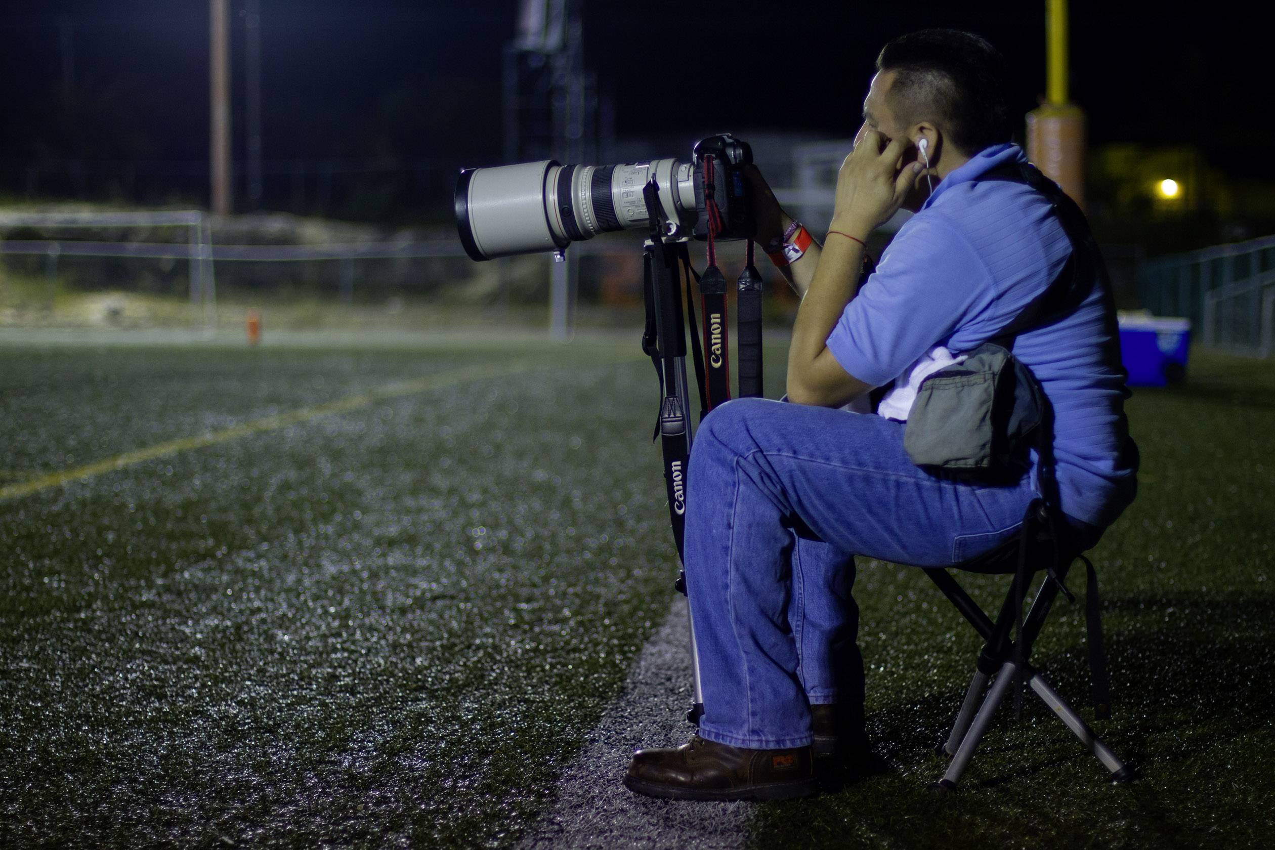 Un photographe est assis sur une chaise au bord du terrain, muni d'un grand appareil photo et d'un objectif, concentré sur l'action en cours.