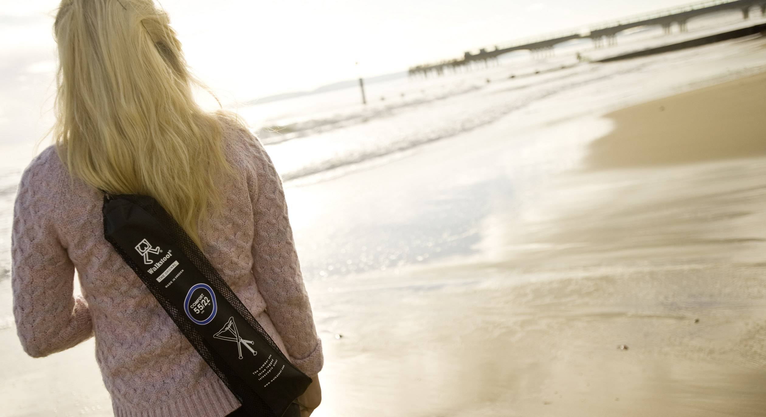 Une personne marche le long de la plage, portant un sac noir avec des logos. Le ciel est nuageux, la mer est calme.