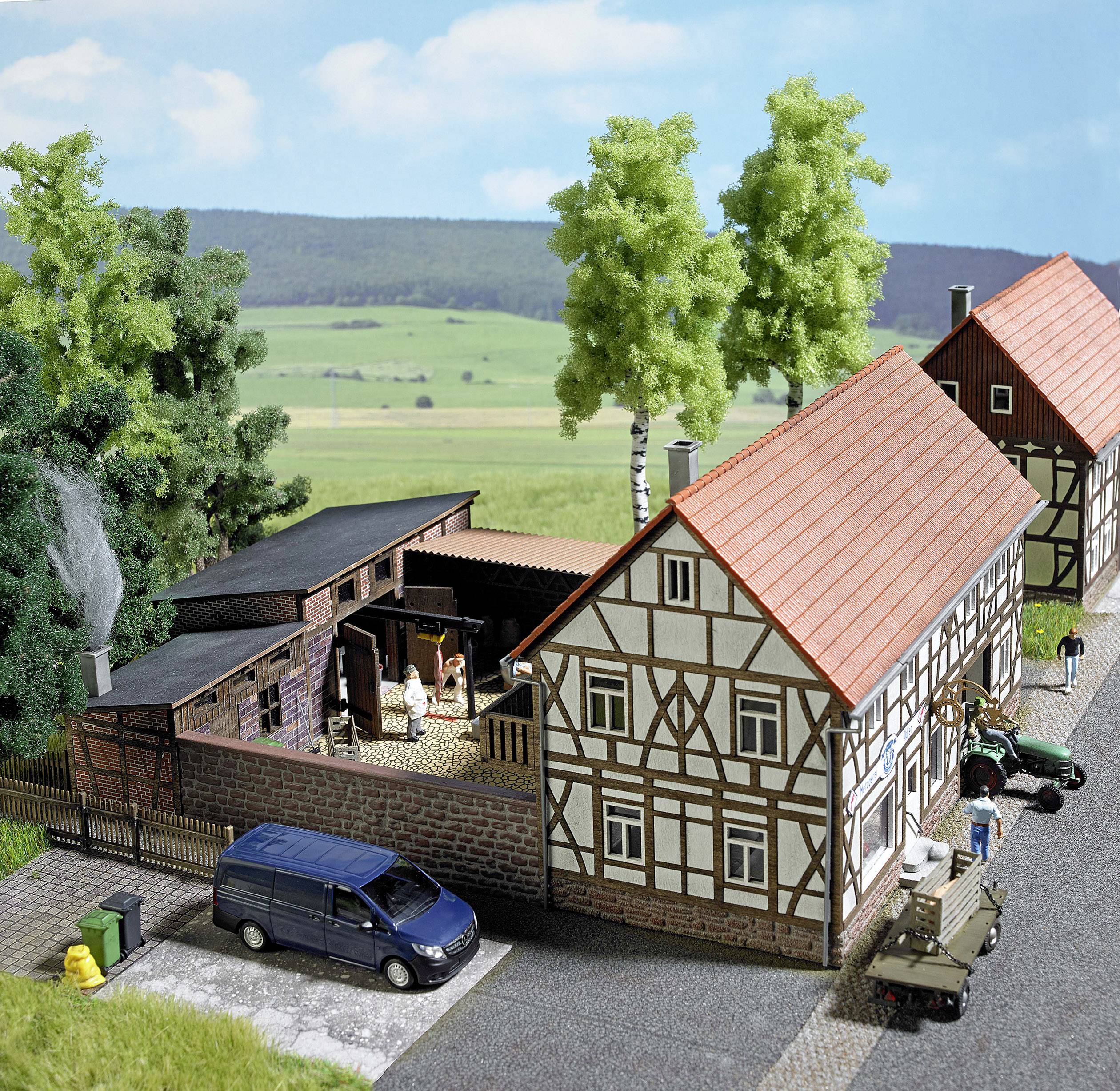 Maquette d'un village avec des maisons à colombages, des arbres et des véhicules. Des personnes sont visibles dans la cour. Paysage rural en arrière-plan.