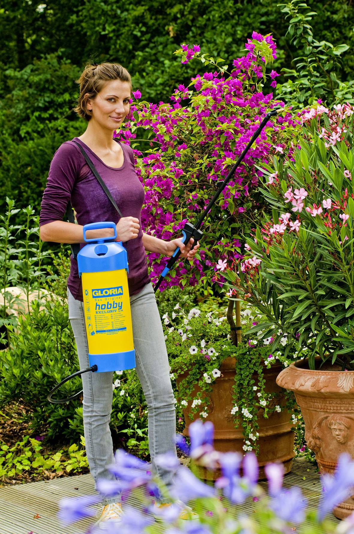 Une femme dans un jardin tient un vaporisateur jaune et un bâton, entourée de plantes en fleurs. Elle porte un pull lilas et un pantalon gris.