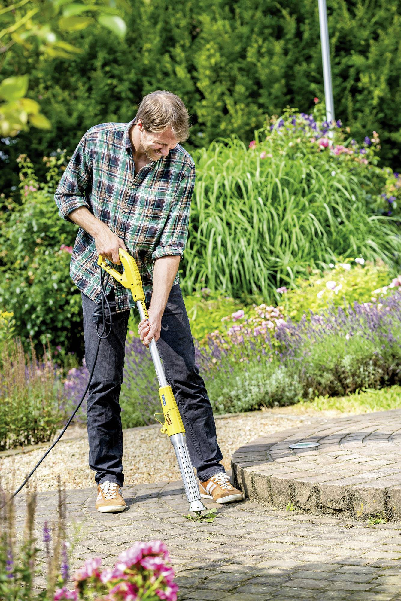 Un homme nettoie un chemin de jardin pavé avec un nettoyeur haute pression jaune. Des parterres de fleurs sont visibles en arrière-plan.