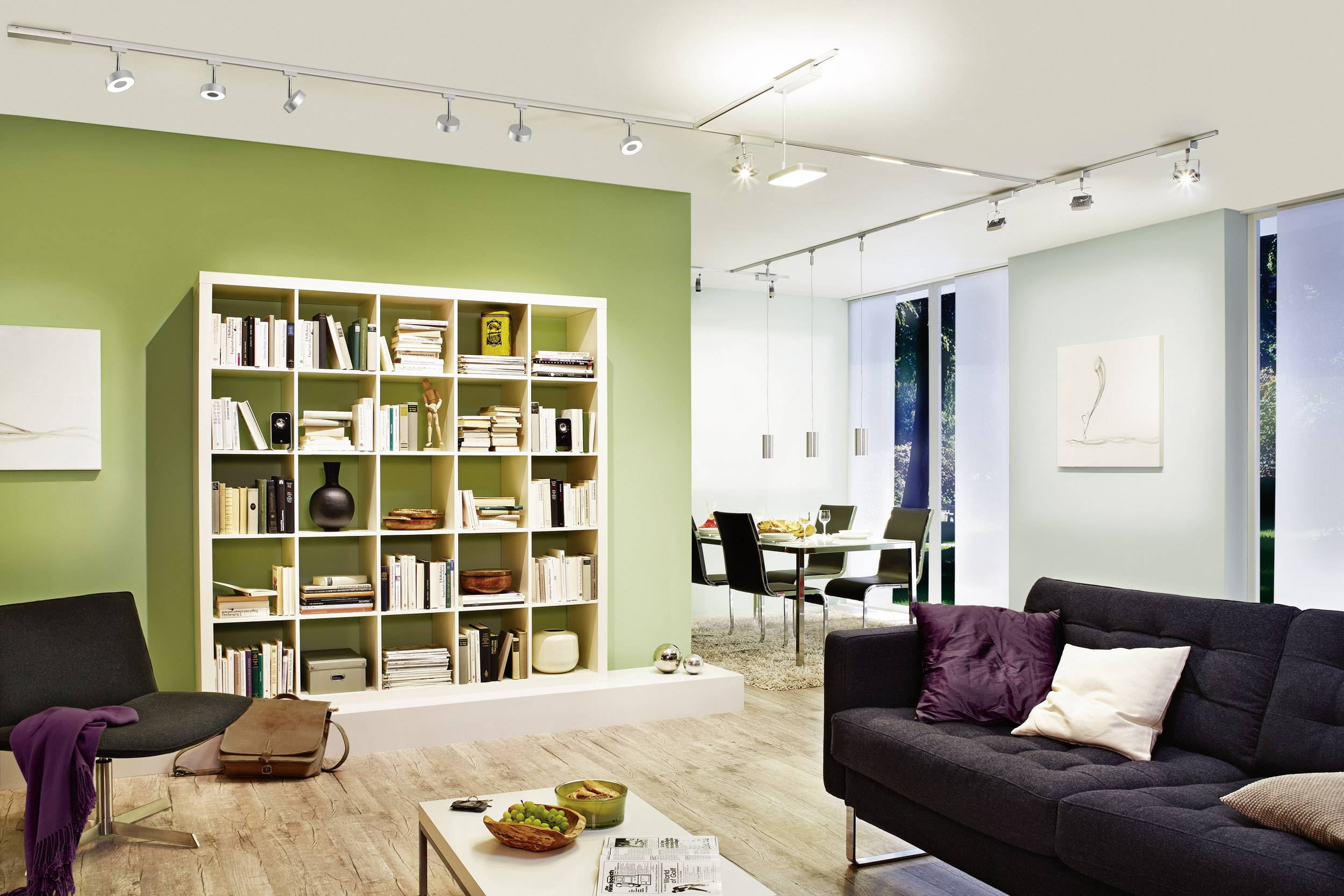 Un salon moderne avec un mur vert et une étagère remplie de livres, un canapé élégant, des luminaires au plafond et un coin repas à l'arrière-plan.