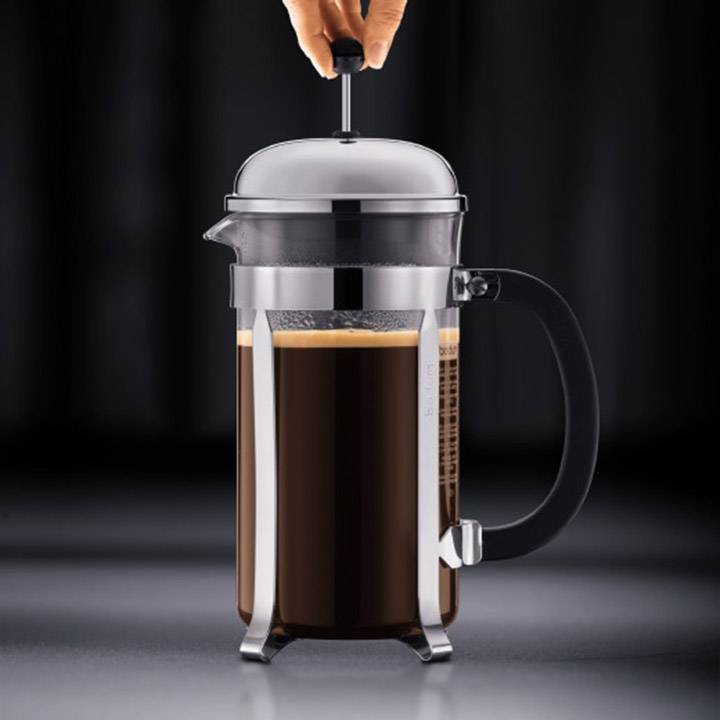 Une main enfonce le piston d'une French Press en verre, alors que du café fraîchement moulu se trouve dans le récipient.
