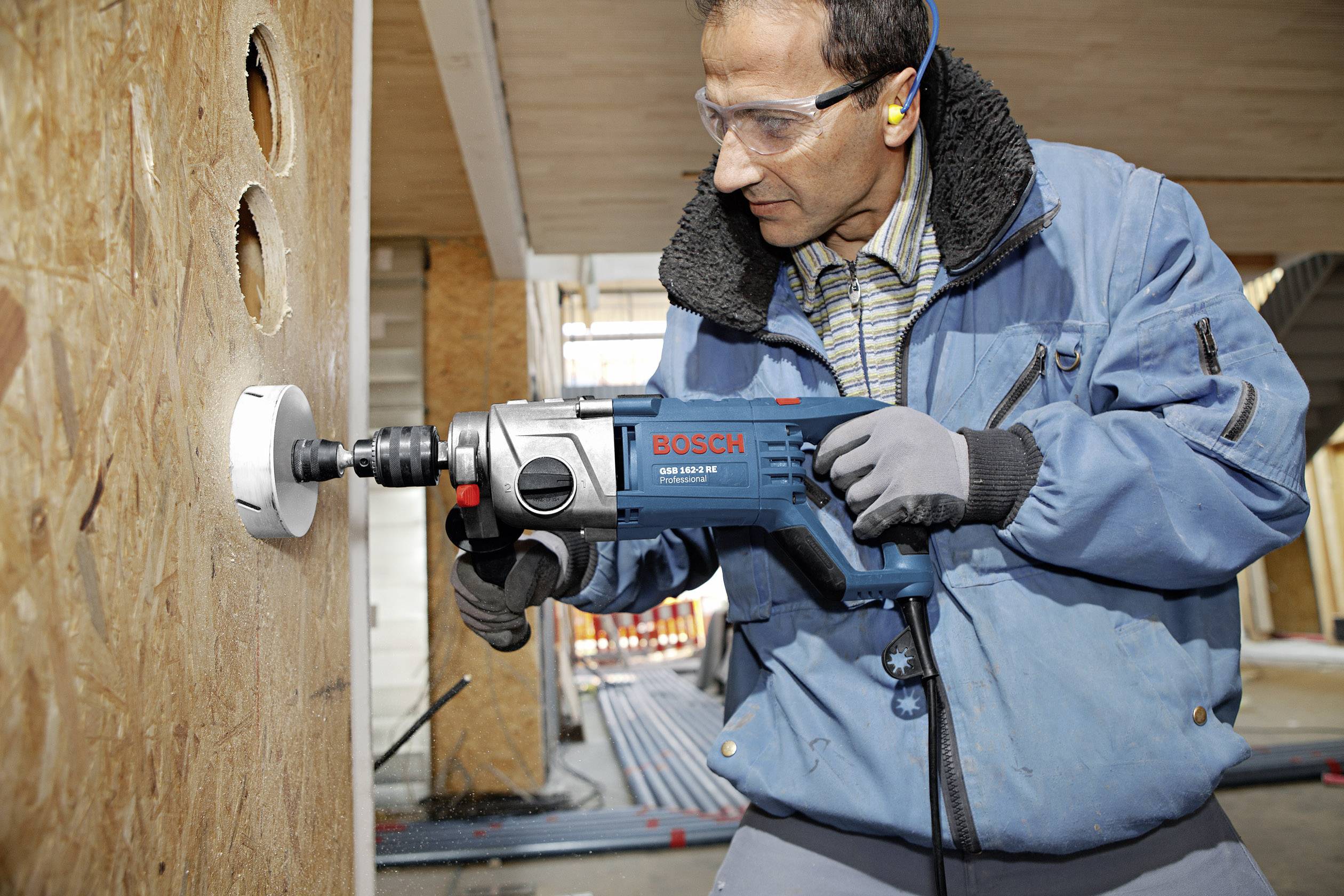 Un homme en équipement de protection fore un grand trou dans une planche de bois à l'aide d'une machine-outil électrique bleue sur un chantier de construction.