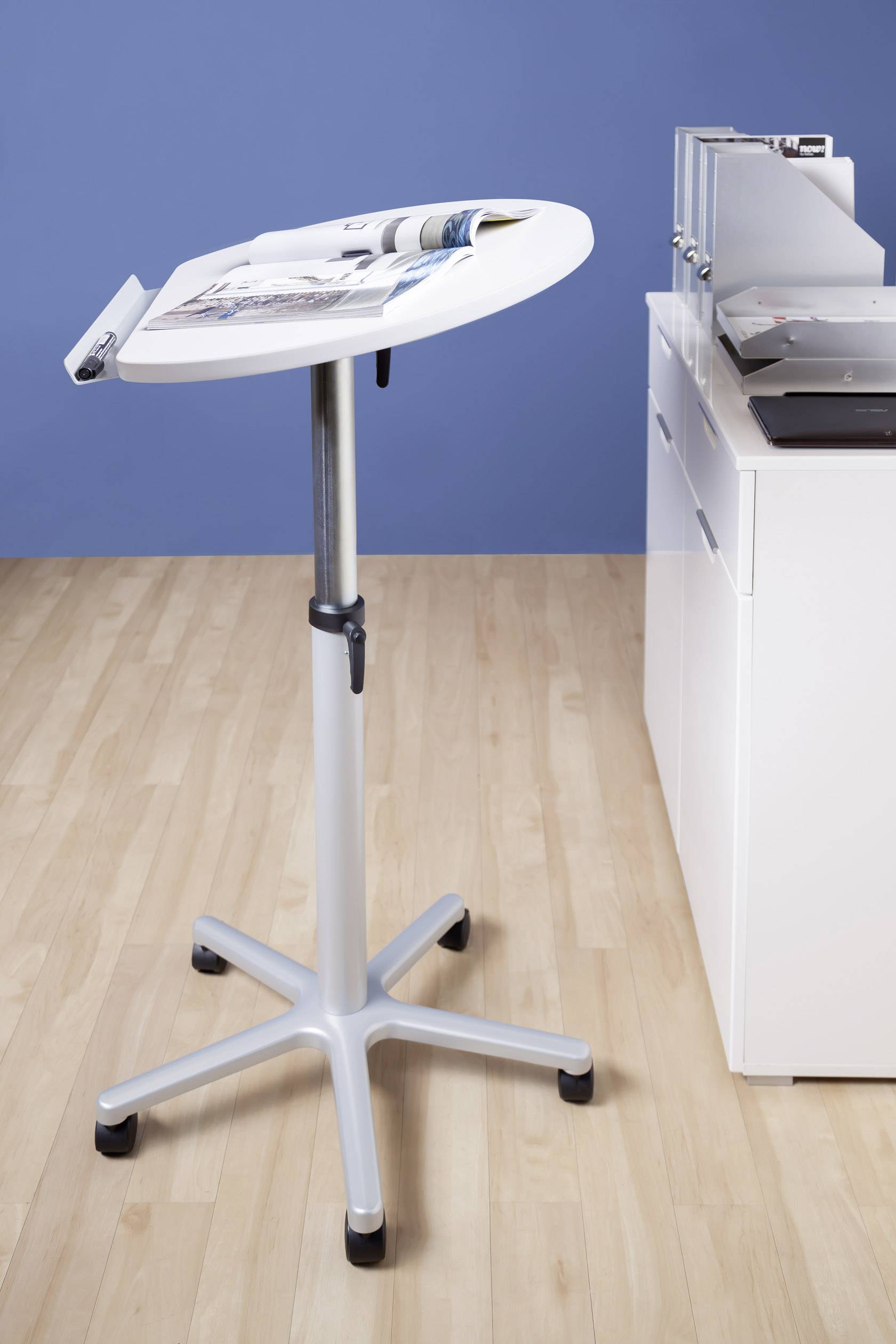 Une table d'appoint réglable en hauteur en métal, montée sur roulettes et chargée de magazines, se trouve à côté d'une armoire de bureau blanche sur un sol en bois.