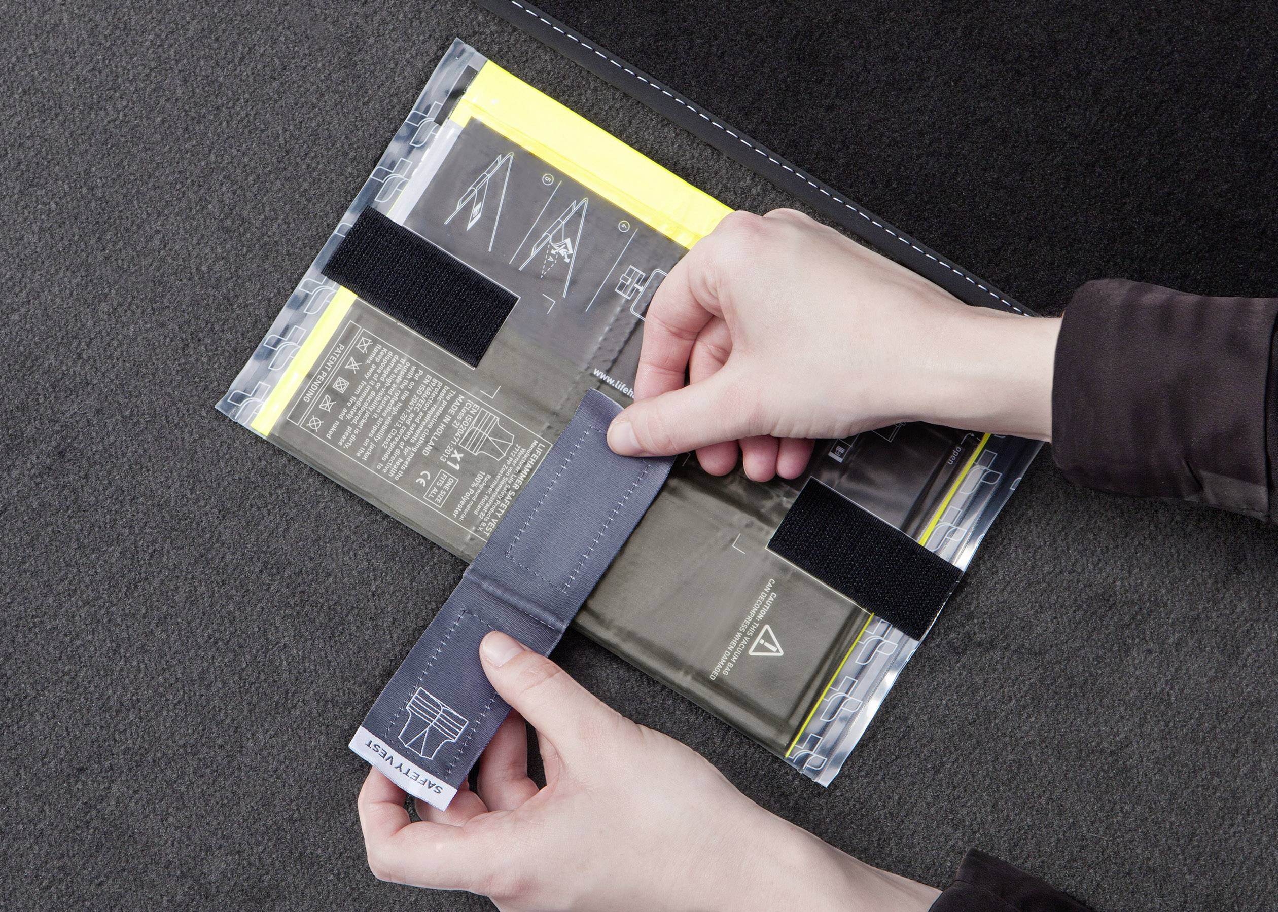 Deux mains ouvrent une pochette noire à plusieurs compartiments, fermée par une bande auto-agrippante. Des documents et des diagrammes sont visibles.