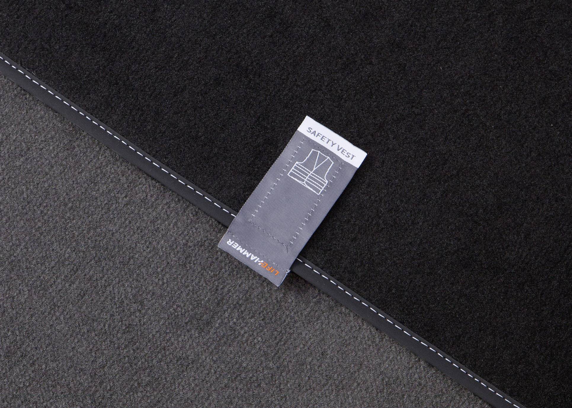 Étiquette portant l'inscription 'SAFETY VEST' sur un tissu sombre. Sur un ruban gris est inscrit 'KOLLEKTIONER'.