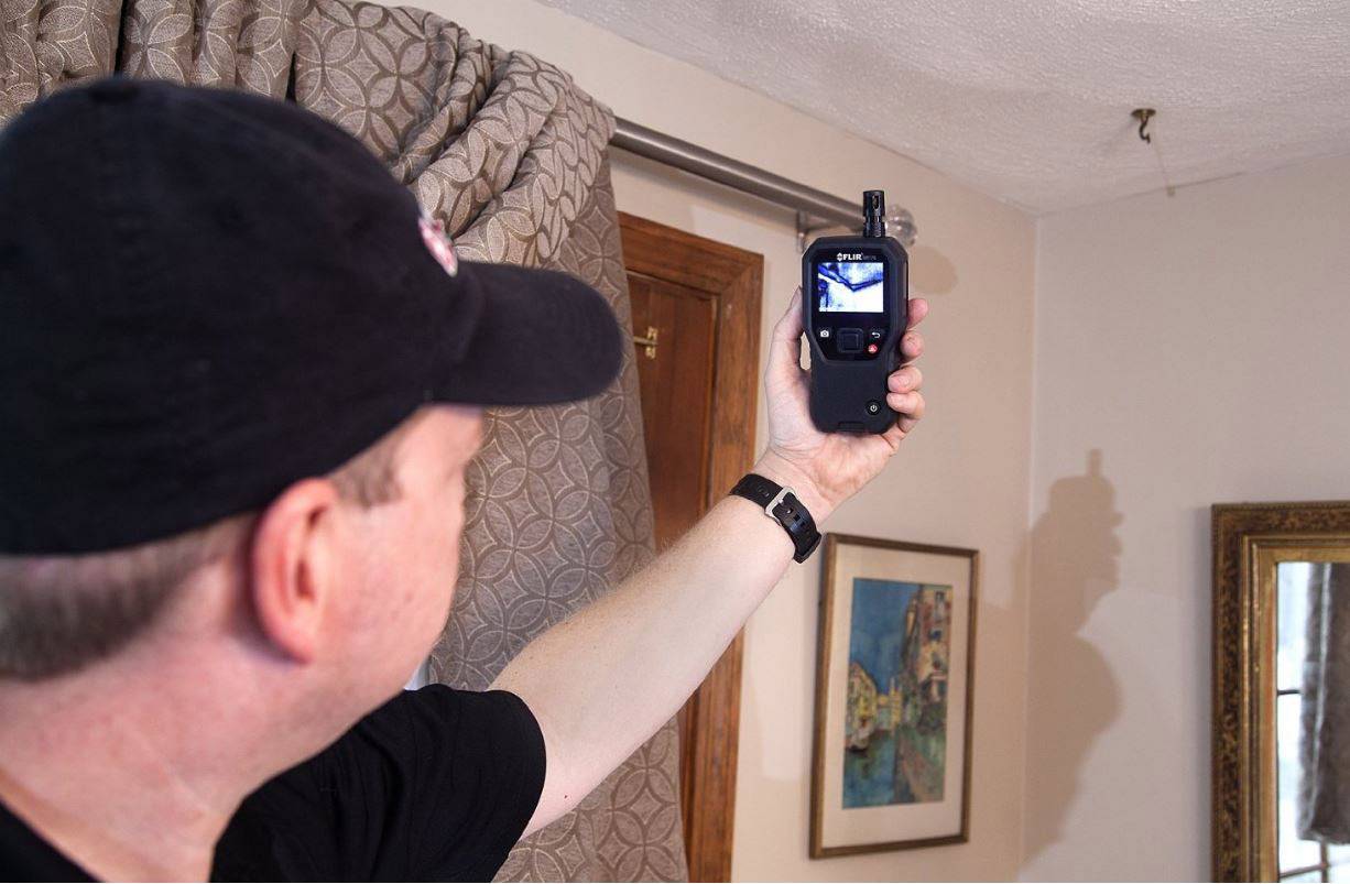 Une personne tient un appareil portable avec un écran, vérifiant des images thermiques dans une pièce. L'arrière-plan montre un rideau et une décoration murale.