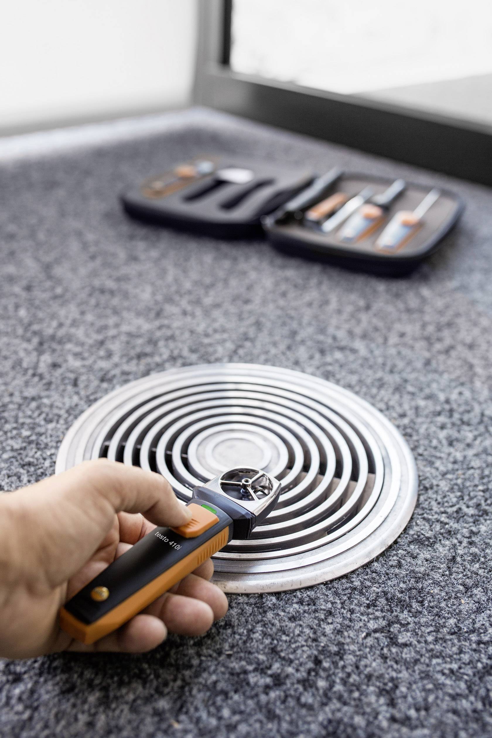 Un homme mesure le débit d'air à une grille de ventilation à l'aide d'un appareil portable. Un coffret à outils ouvert est visible en arrière-plan.