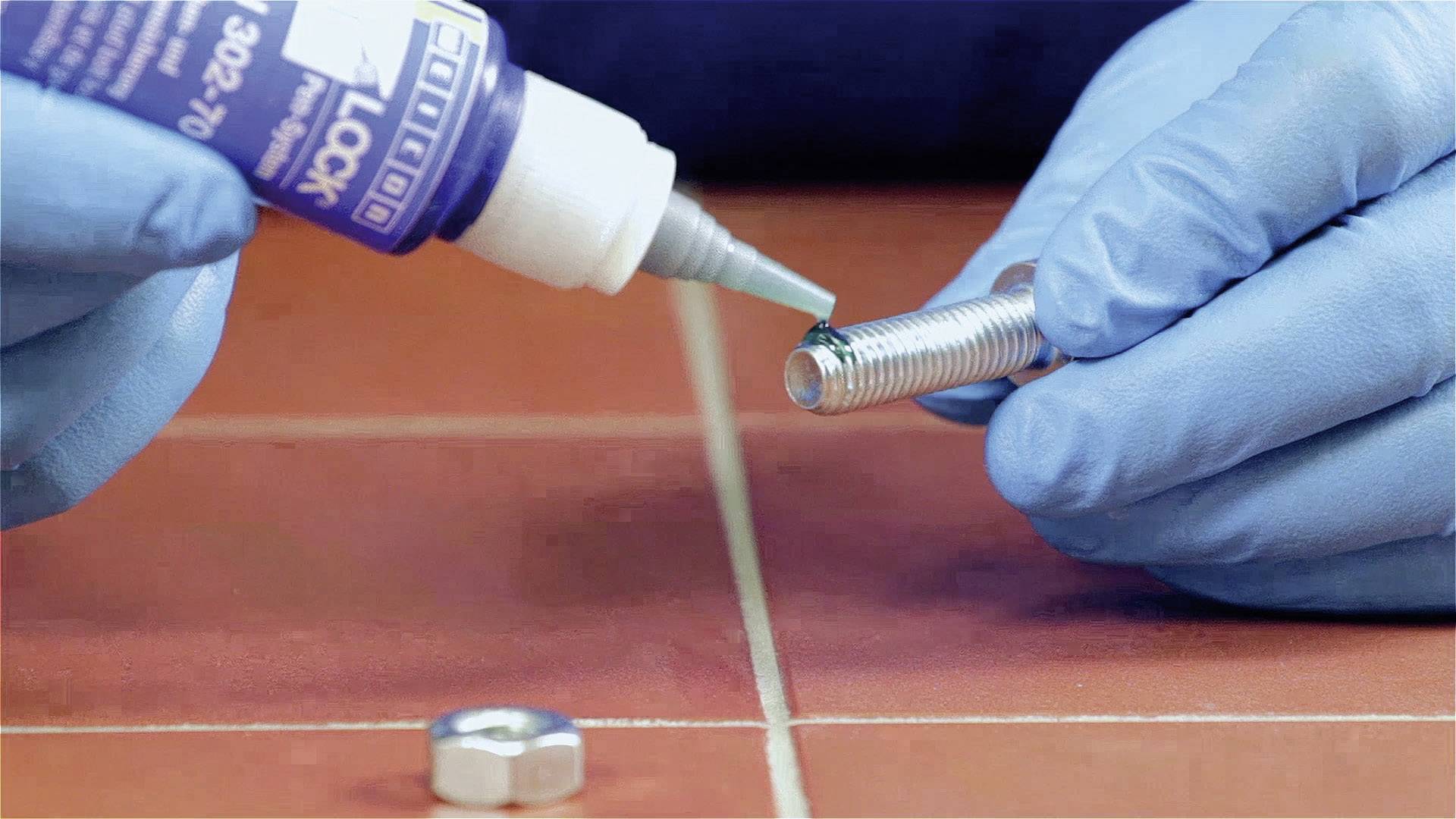 Une personne portant des gants bleus applique de la colle sur un filetage métallique pour le sécuriser.