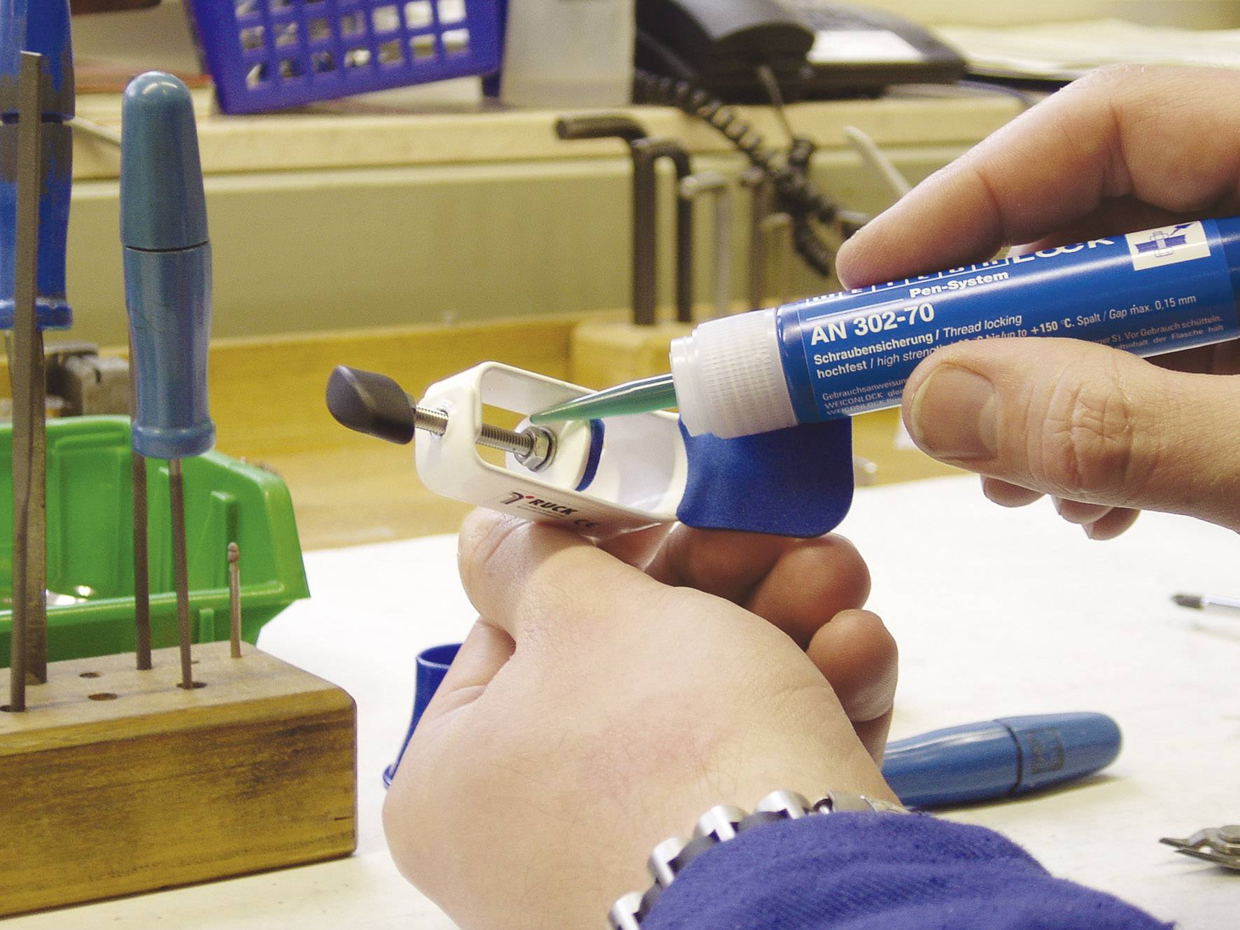 Une personne applique de la colle bleue sur un boulon fileté dans un atelier pour le sécuriser. Des outils sont visibles en arrière-plan.