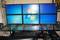 Un poste de travail avec six écrans affiche le logo Windows sur un fond bleu. Un clavier et une souris sont posés sur le bureau.