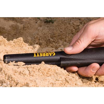 Une main tient un détecteur de métaux, partiellement enfoui dans le sable. L'appareil est noir avec l'inscription jaune 'Garrett'.