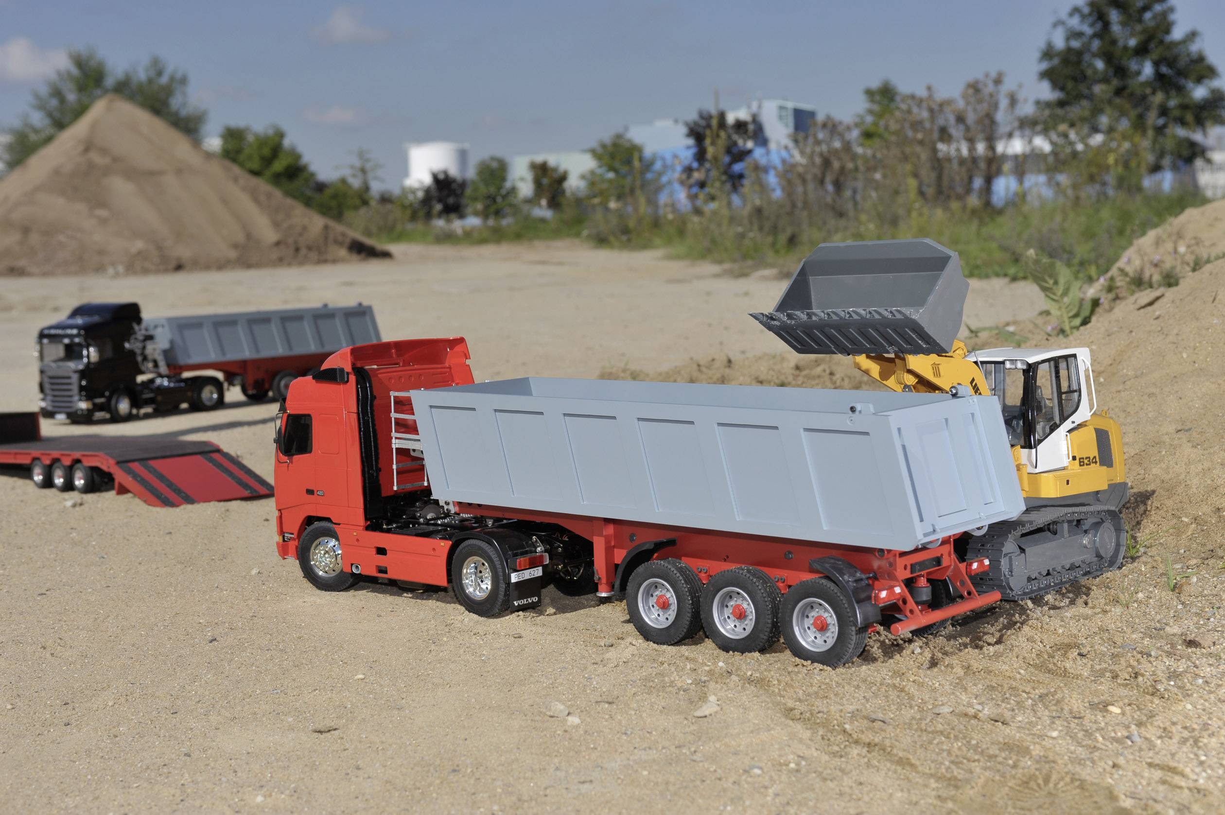 Un modèle de camion jouet rouge avec remorque est chargé par un pelleteuse gris, placé sur un chantier sablonneux.