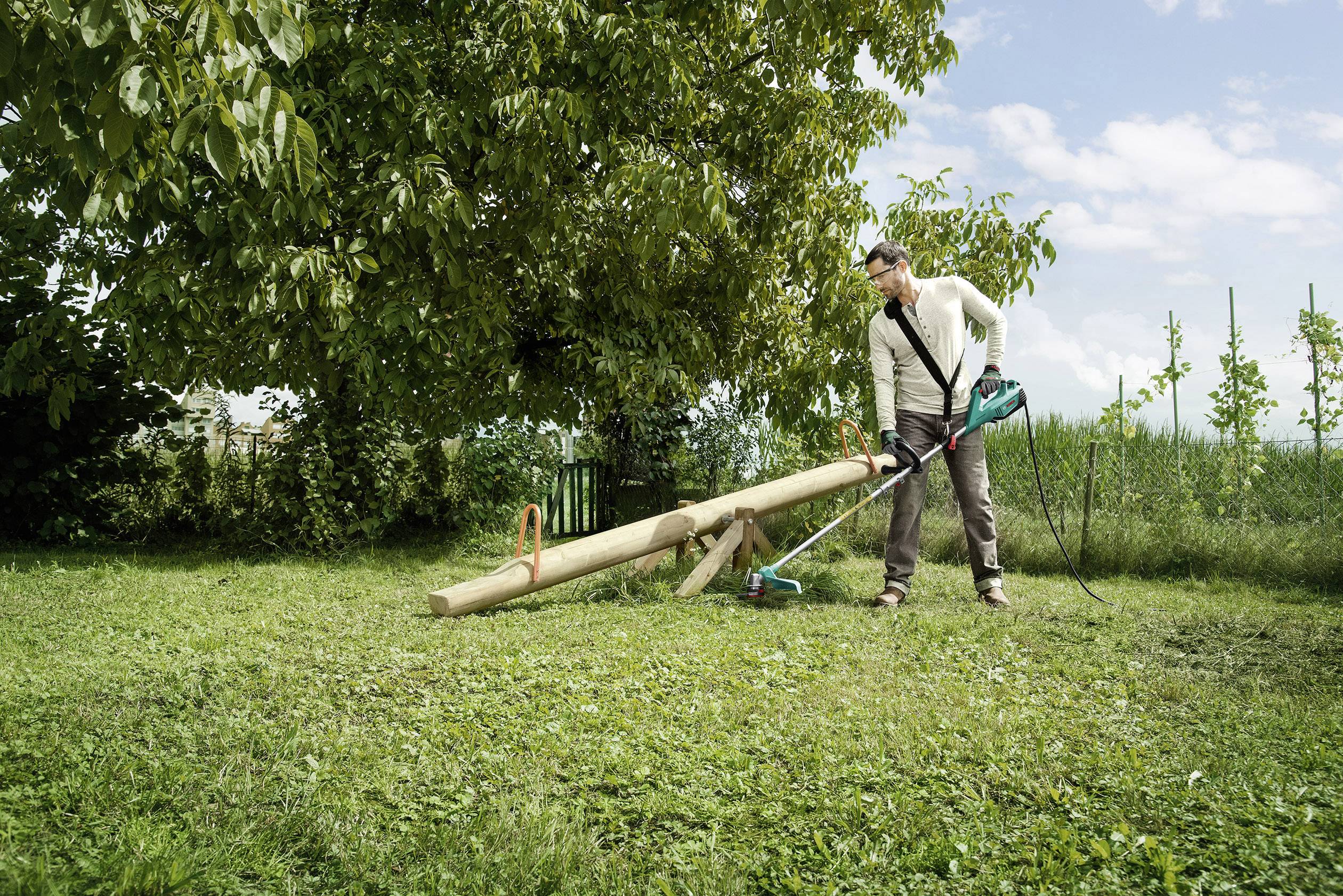 Un homme tond la pelouse avec un coupe-herbe électrique dans un jardin. Des arbres et une clôture sont visibles en arrière-plan.