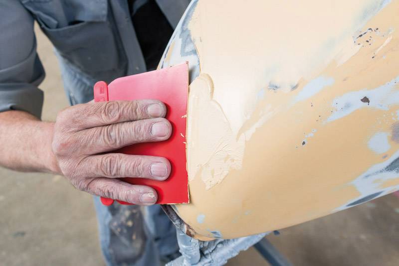 Un homme ponce une surface beige vernie avec un outil rouge. La surface présente des traces d'usure et de réparation.
