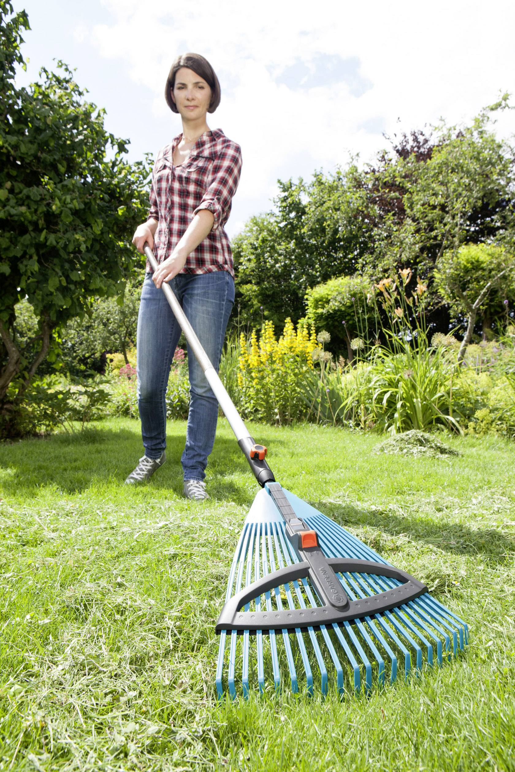 Une personne râtisse de l'herbe avec un râteau bleu dans un jardin. Des fleurs et des buissons sont visibles en arrière-plan. C'est une journée ensoleillée.