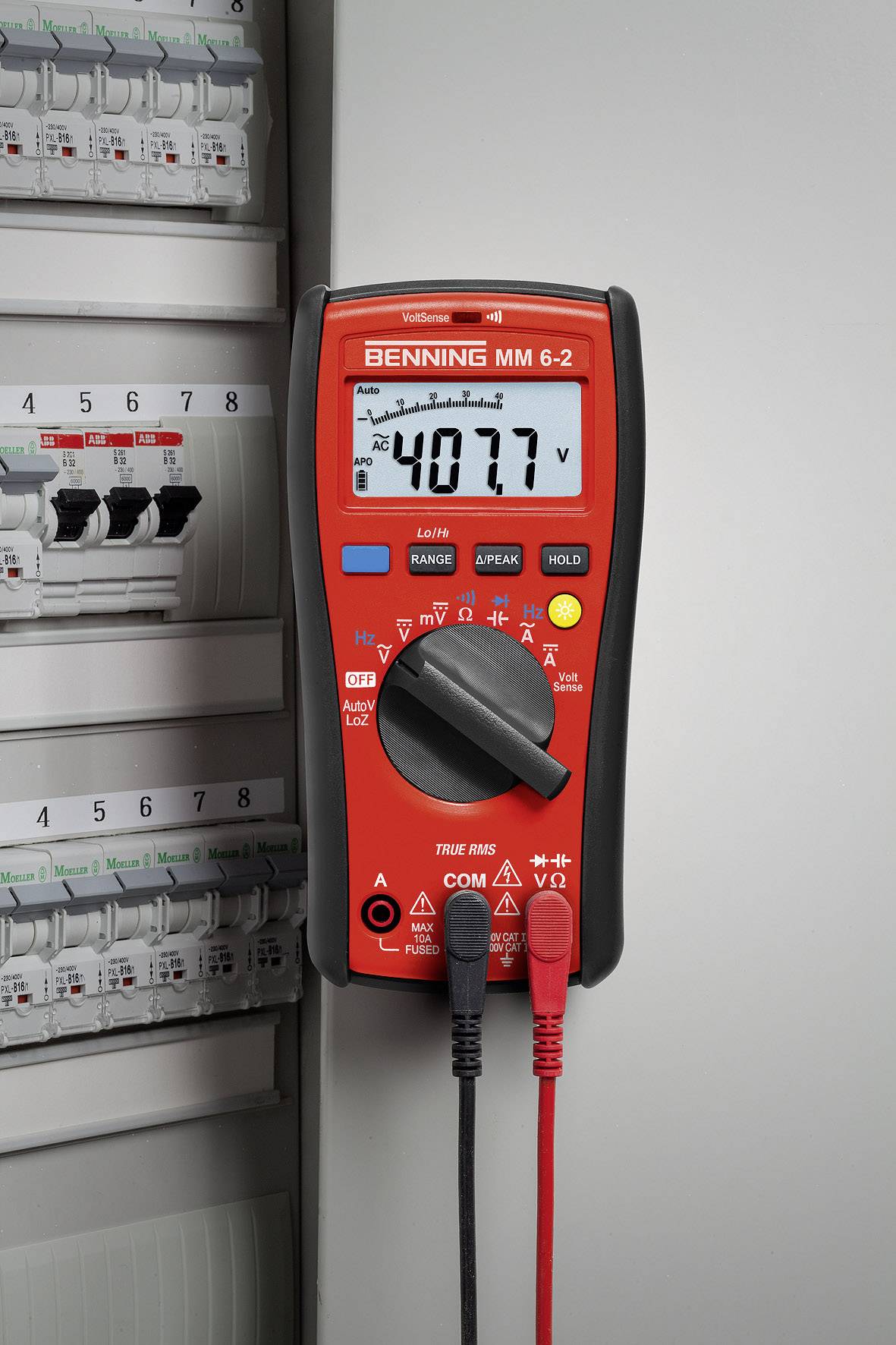 Un multimètre rouge affiche une tension de 40,77 sur un écran électronique et est connecté à un tableau électrique.