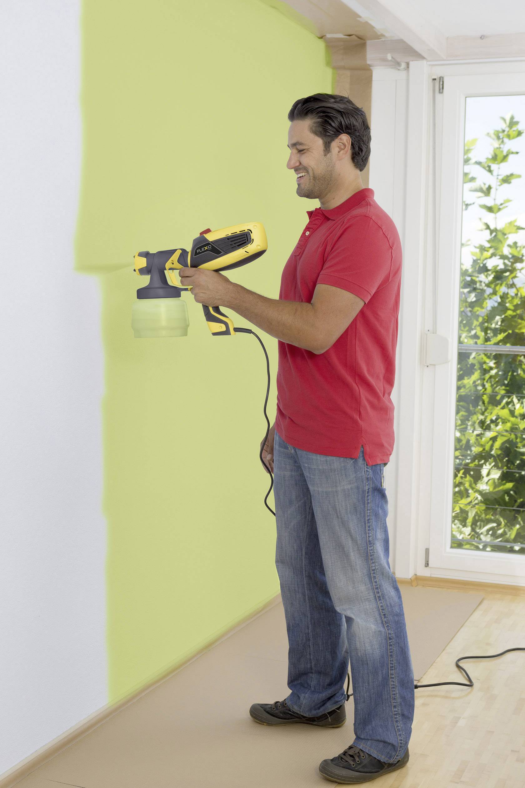 Un homme portant une chemise rouge peint un mur en vert à l'aide d'un pistolet à peinture. Il se trouve dans une pièce avec de grandes fenêtres.