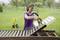 Une femme peint un banc en bois avec un pistolet à peinture dans le jardin. Des arbres et une prairie verte sont visibles en arrière-plan.