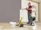 Un homme peint un mur dans une pièce à l'aide d'un pulvérisateur de peinture. Devant lui se trouve un seau de peinture. Le pulvérisateur est jaune.
