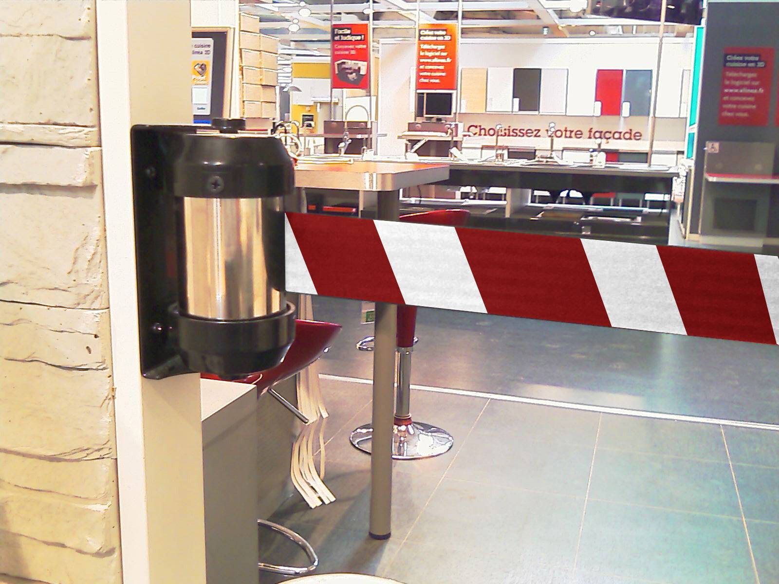 Un ruban de signalisation à rayures rouges et blanches bloque l'accès à une zone de cuisine dans un magasin. Des tables et des chaises sont visibles en arrière-plan.