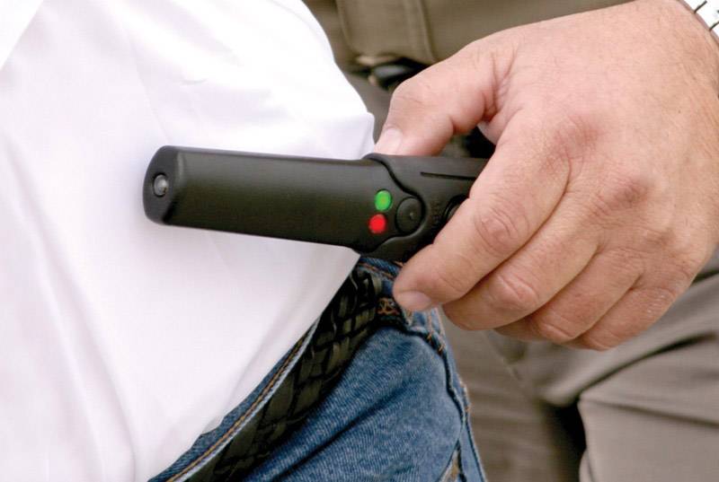 Une personne tient un détecteur de métaux portable au-dessus de la poche d'un pantalon d'une autre personne. Le détecteur affiche des voyants verts et rouges.