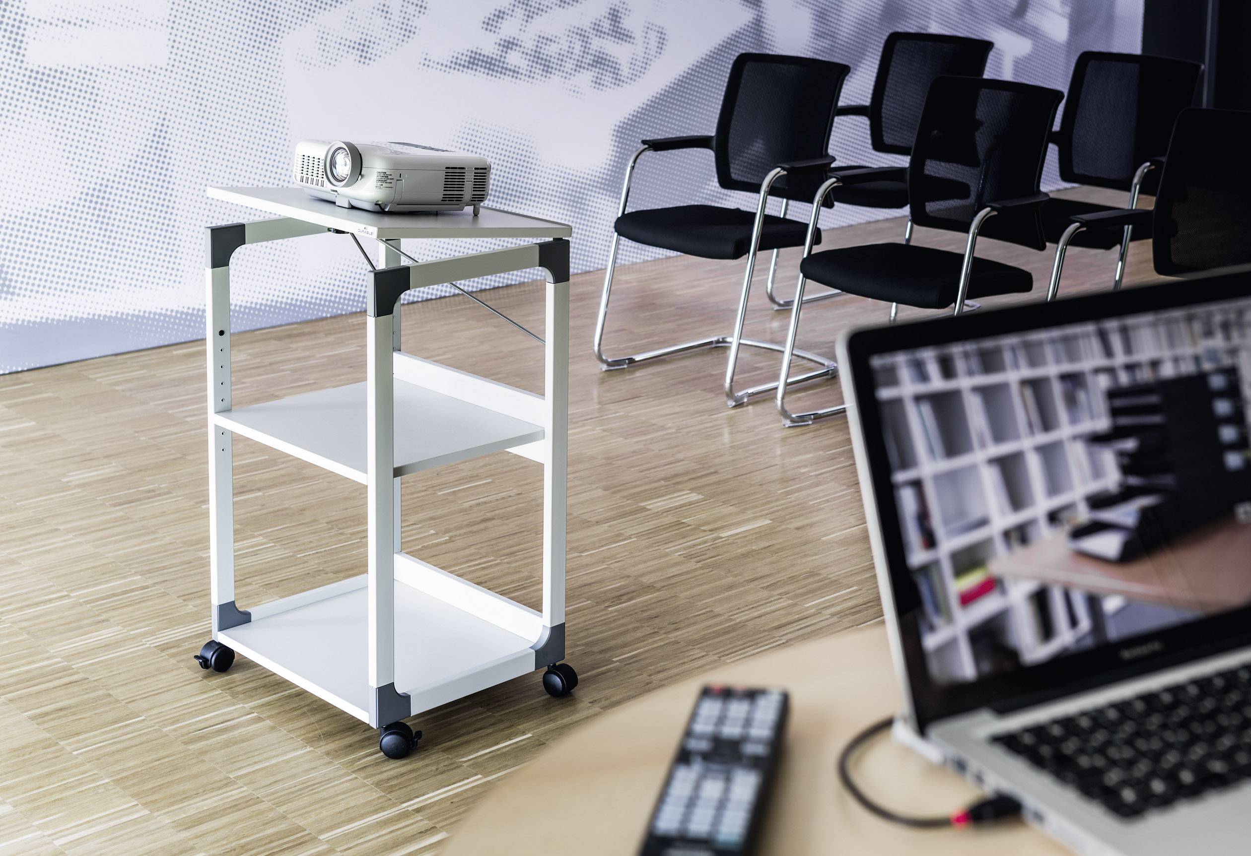 Un chariot de présentation avec un projecteur est au premier plan. À l'arrière-plan, des chaises vides sont visibles. Un ordinateur portable affiche une image floue.