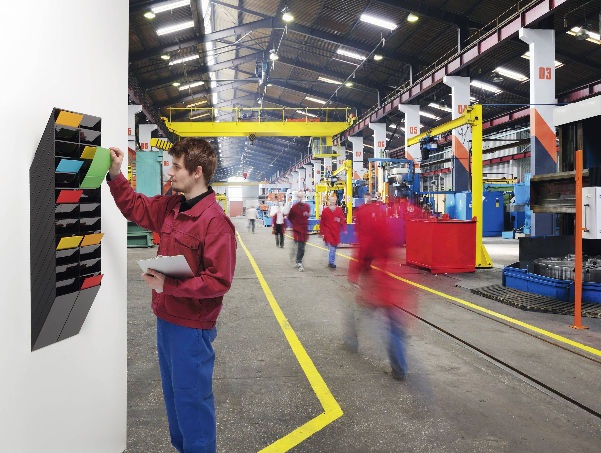 Une personne en tenue de travail vérifie des documents sur une étagère murale dans une grande halle d'usine animée, avec d'autres ouvriers en arrière-plan.