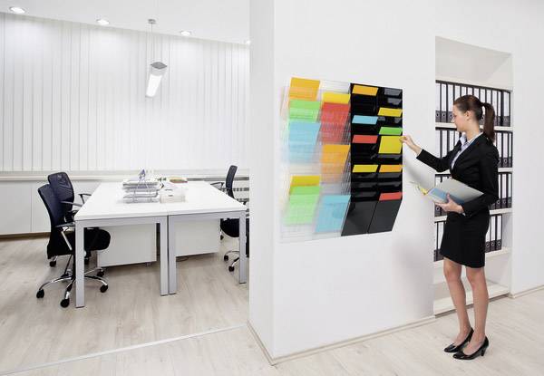 Une femme en tenue professionnelle se tient devant des étagères murales dans un bureau moderne, tenant des classeurs et retirant un document.