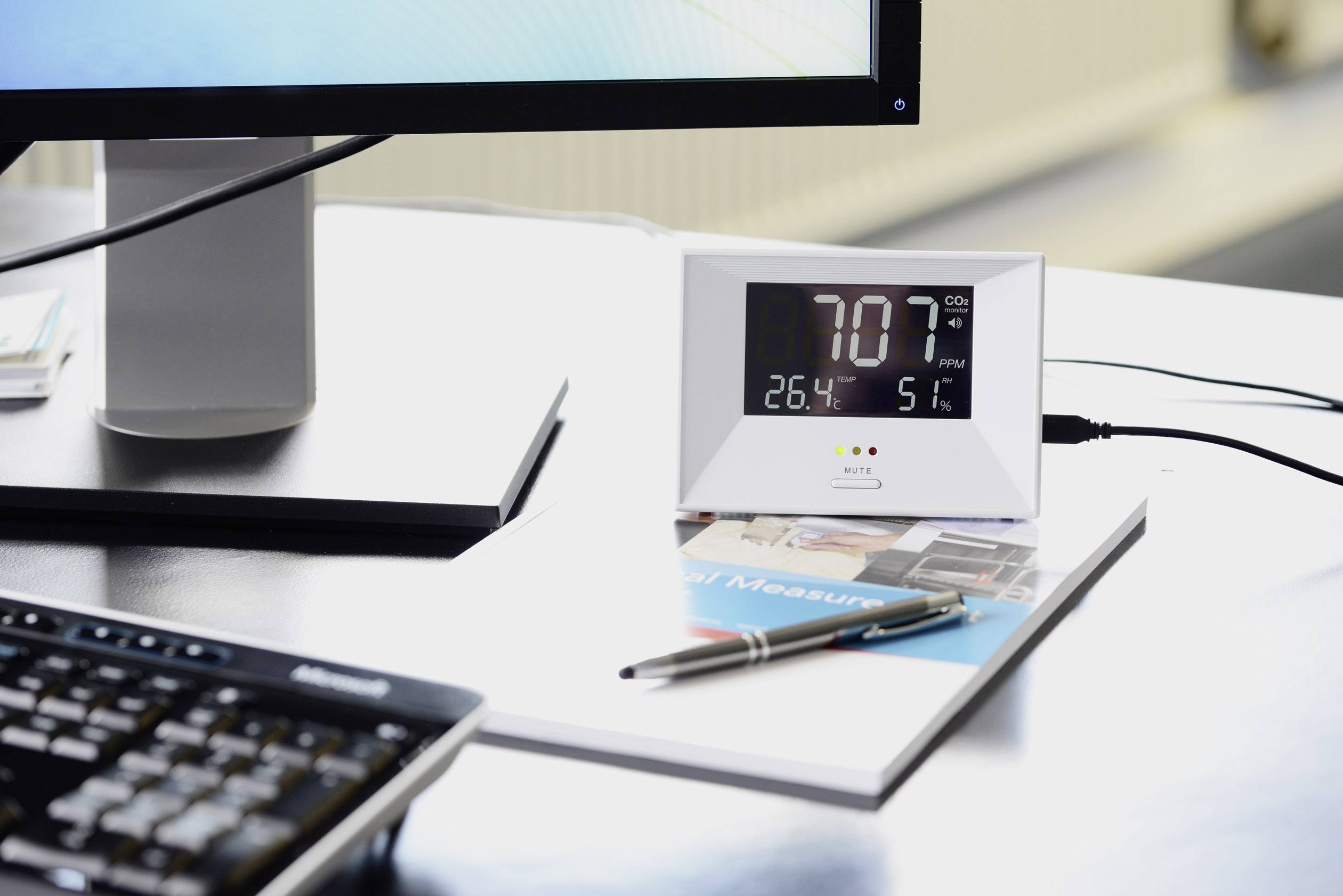 Une table avec un appareil de mesure du CO2 à côté d'un écran d'ordinateur. L'appareil affiche '707 ppm CO2', '26,4°C' et '51% d'humidité'.