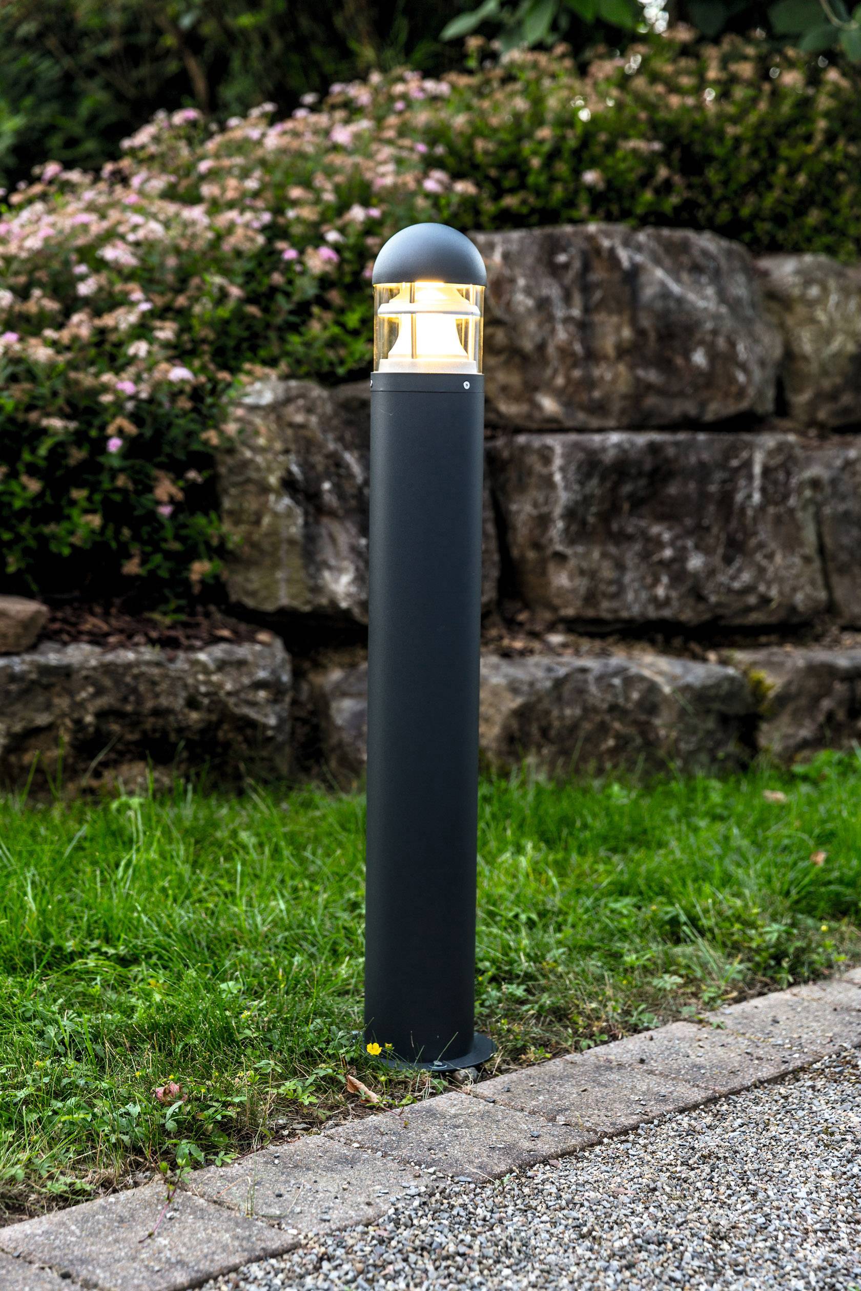 Une lampe de jardin est posée sur une pelouse à côté d'un chemin pavé, avec un mur de pierre et des arbustes en fleurs en arrière-plan.