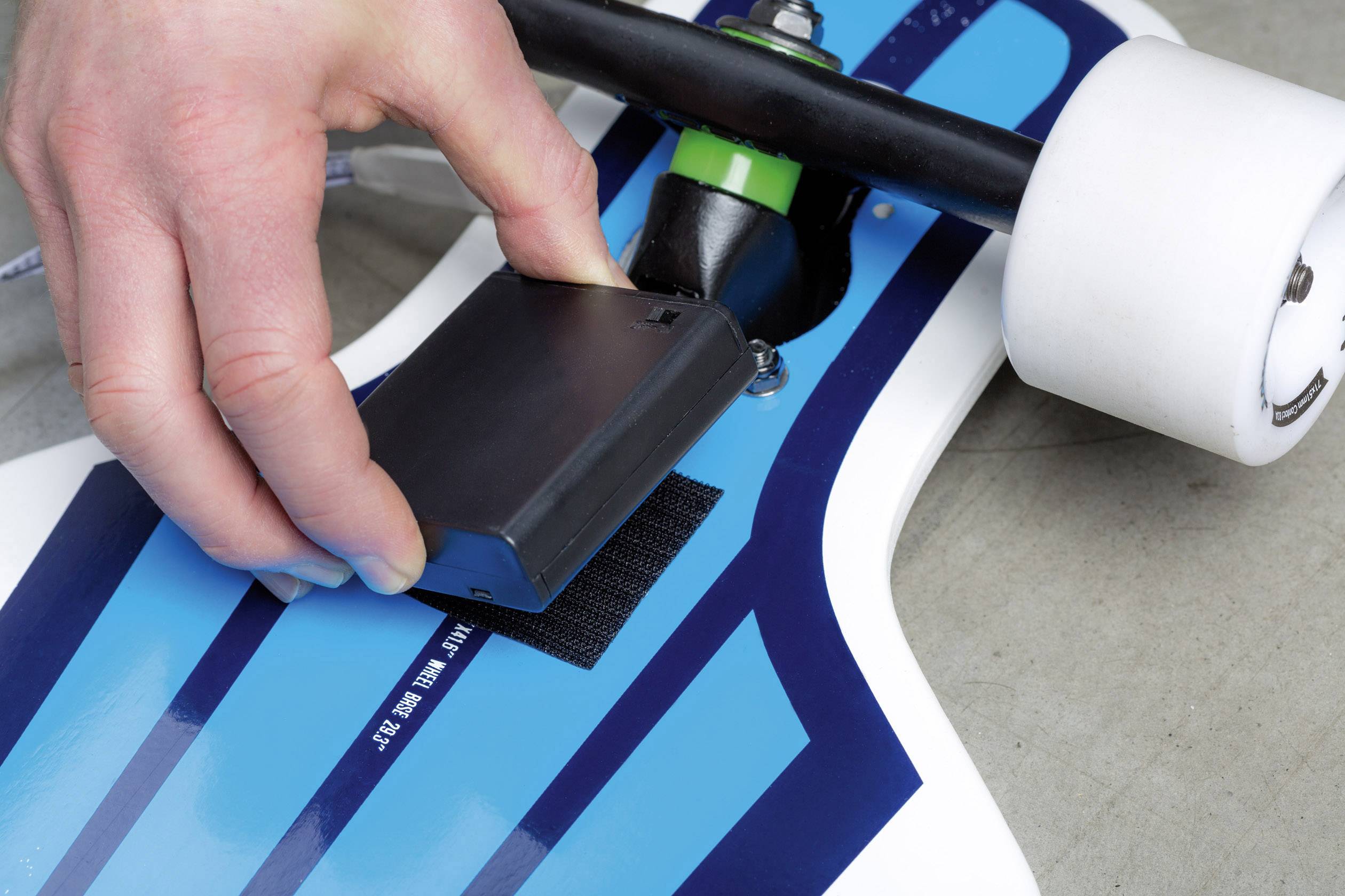 Une personne fixe un objet rectangulaire sous un longboard bleu. Le focus est mis sur la main et l'objet.