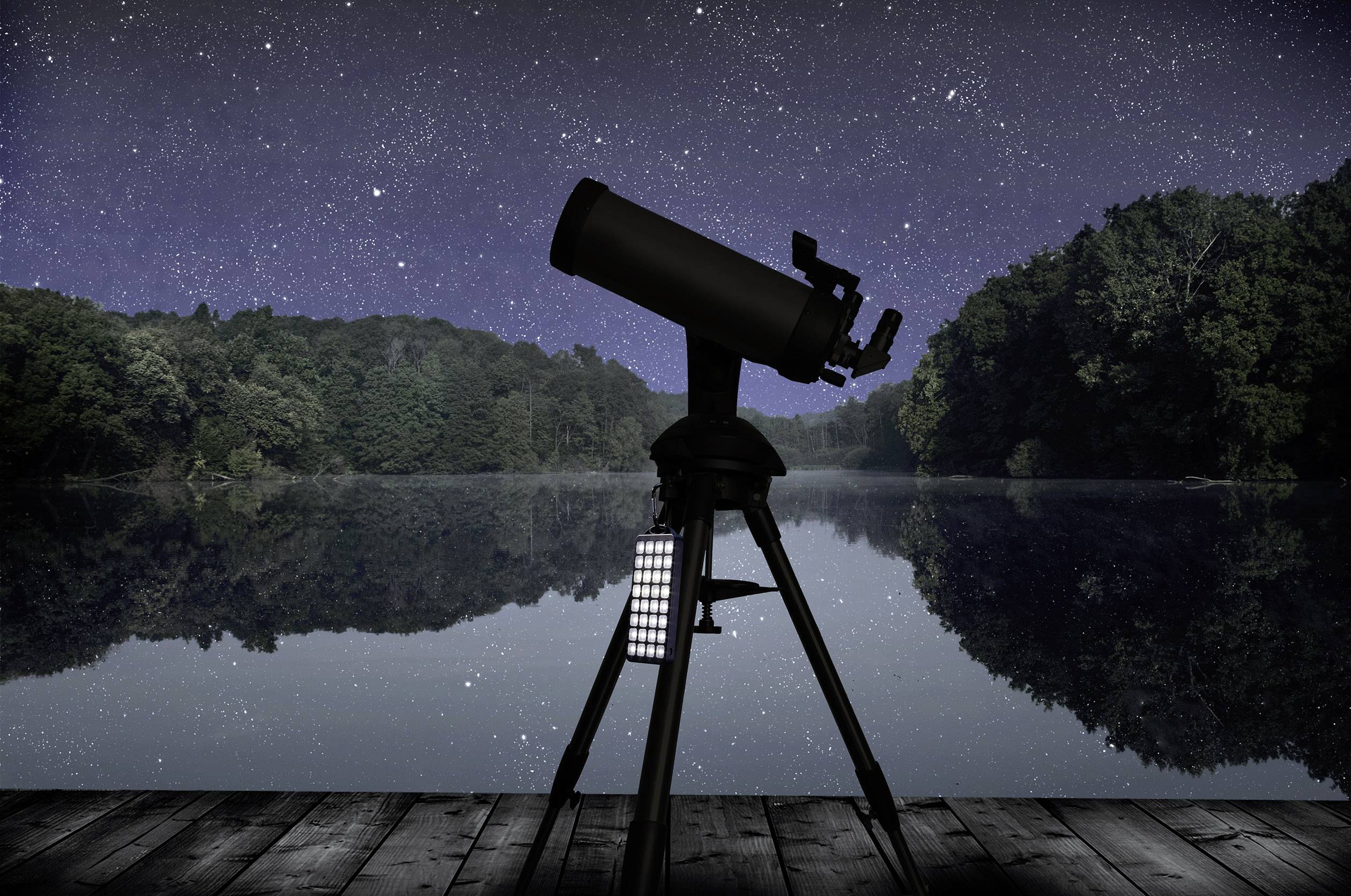 Un télescope est posé sur une terrasse en bois et observe un ciel nocturne étoilé au-dessus d'un lac paisible, entouré d'arbres.