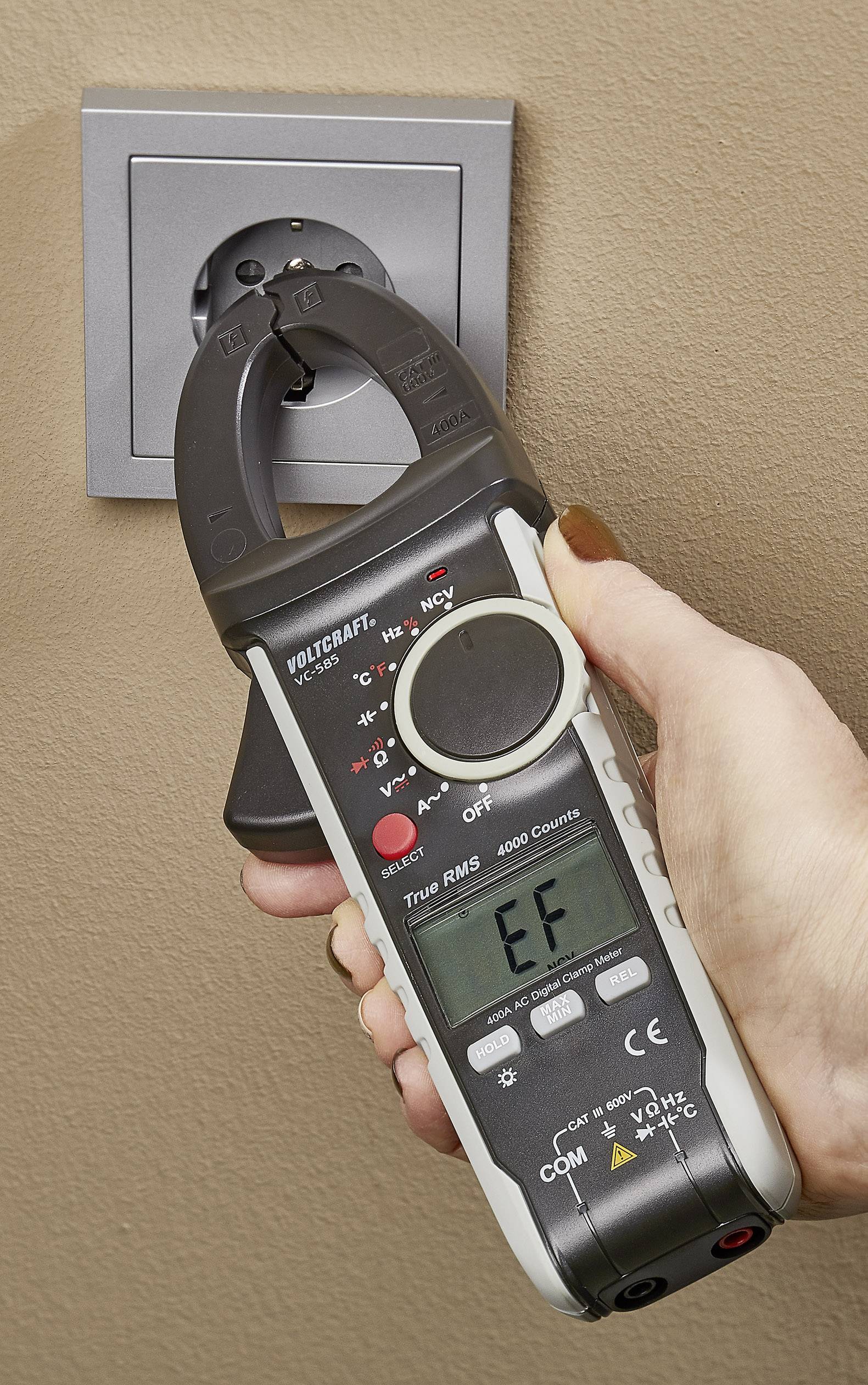 Une main tient un multimètre mesurant une prise électrique. L'affichage indique 'EF'.