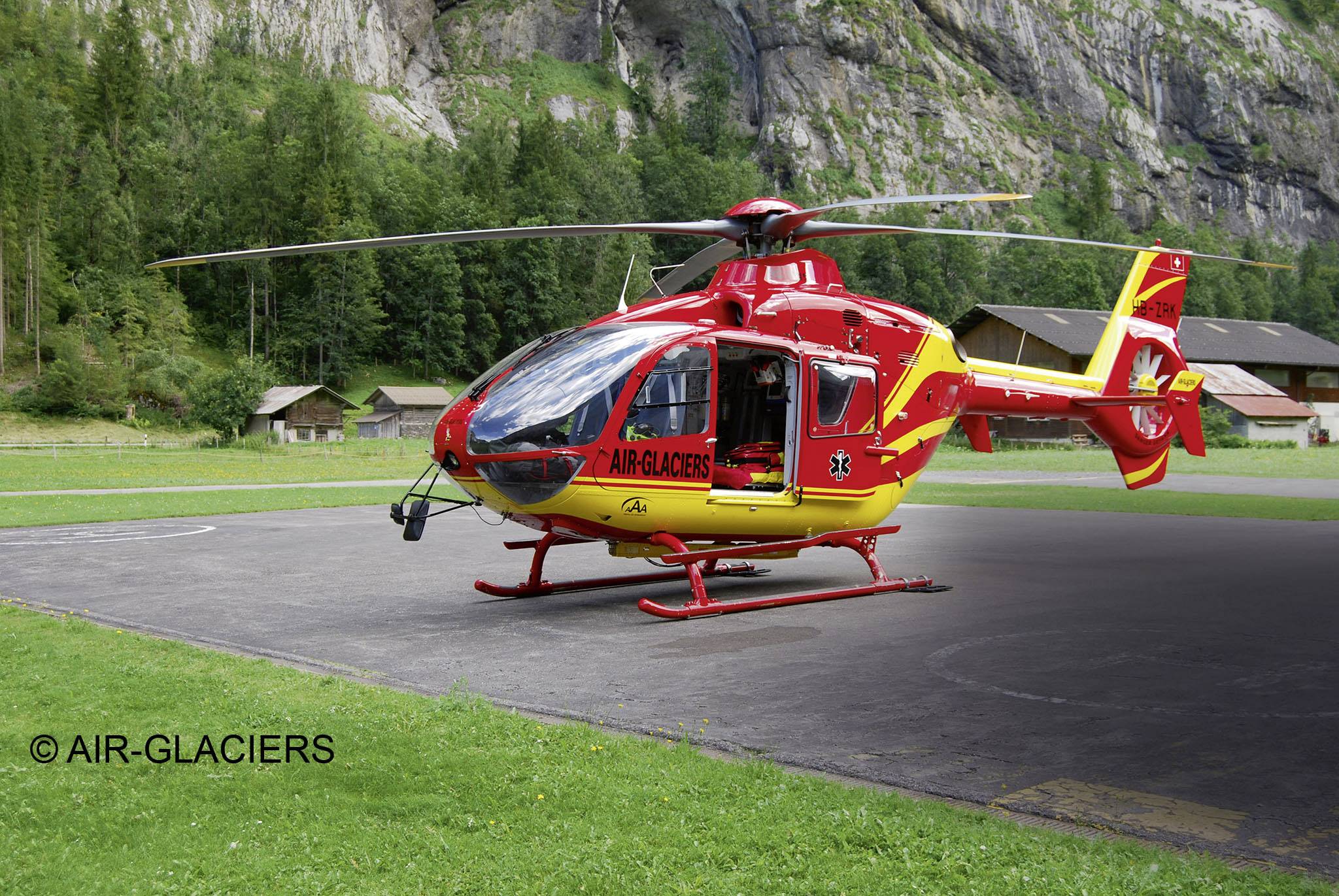 Un hélicoptère de secours rouge avec des accents jaunes de la 'Air-Glaciers' est stationné sur une aire d'atterrissage devant un paysage montagneux boisé.