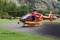 Un hélicoptère de secours rouge avec des accents jaunes de la 'Air-Glaciers' est stationné sur une aire d'atterrissage devant un paysage montagneux boisé.