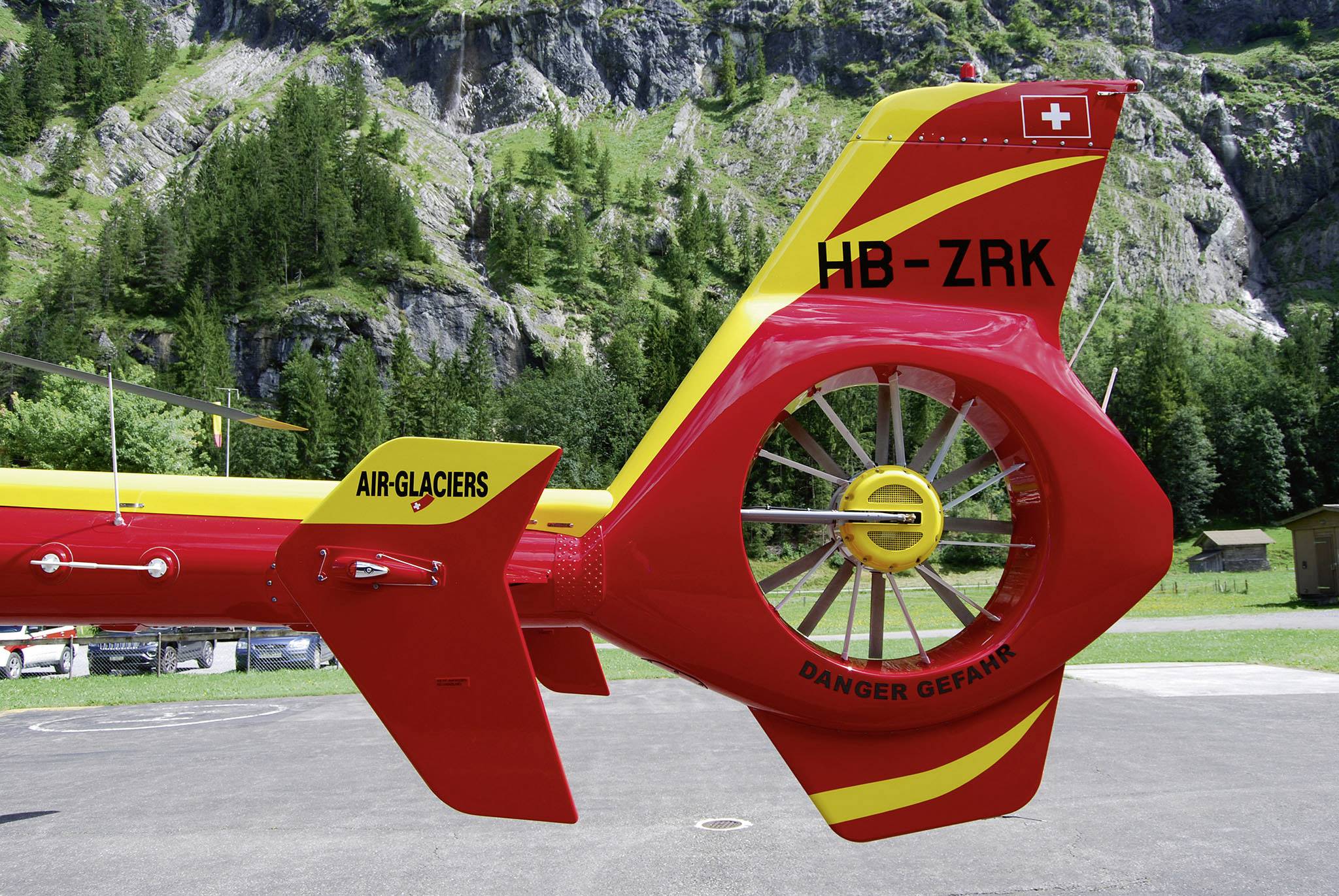 Rotor arrière d'un hélicoptère rouge sur une aire d'atterrissage, portant les inscriptions 'AIR-GLACIERS' et 'HB-ZRK'. Des montagnes et une forêt sont visibles en arrière-plan.