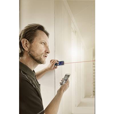 Un homme mesure la distance jusqu'à un mur dans une pièce lumineuse à l'aide d'un télémètre laser. Il tient un smartphone dans son autre main.