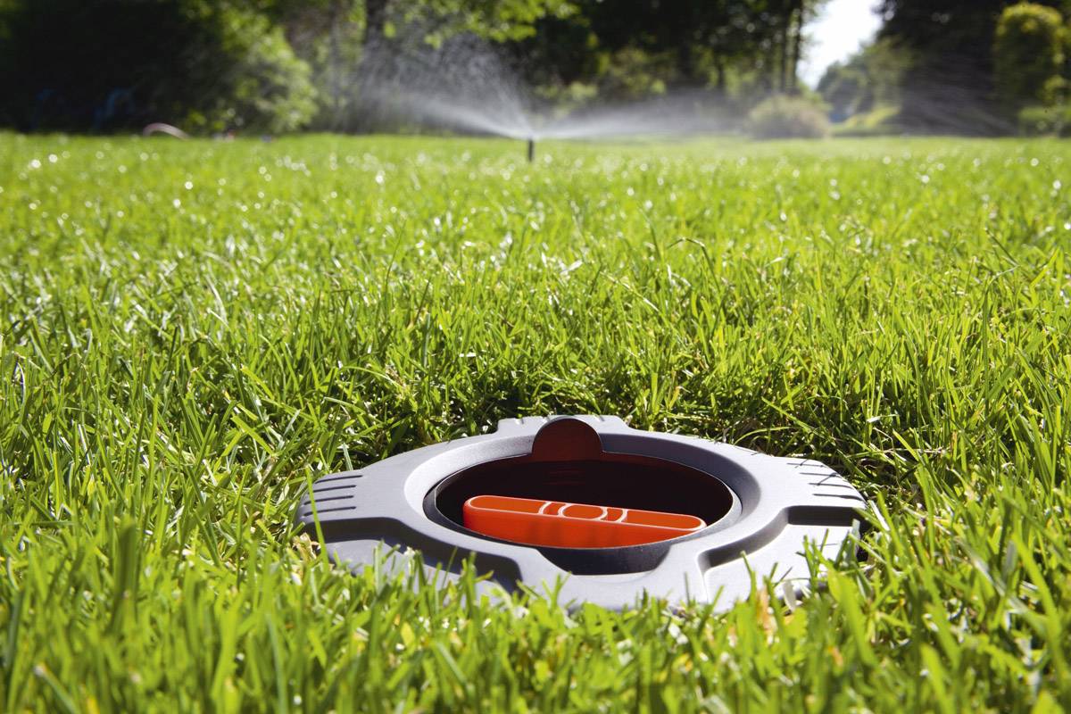 Un gros plan d'un arroseur automatique dans l'herbe, prêt à fonctionner. À l'arrière-plan, de l'eau est projetée sur un pré verdoyant.