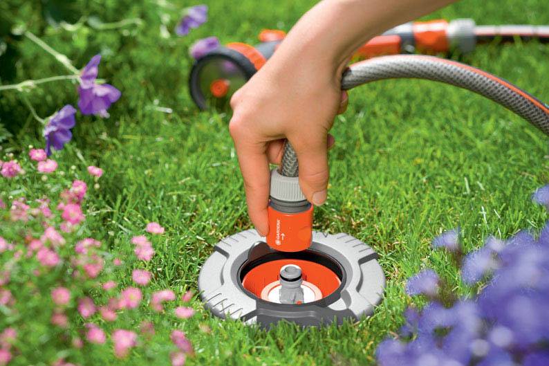 Une main insère un tuyau d'arrosage dans un raccordement au sol dans l'herbe, entouré de fleurs colorées.