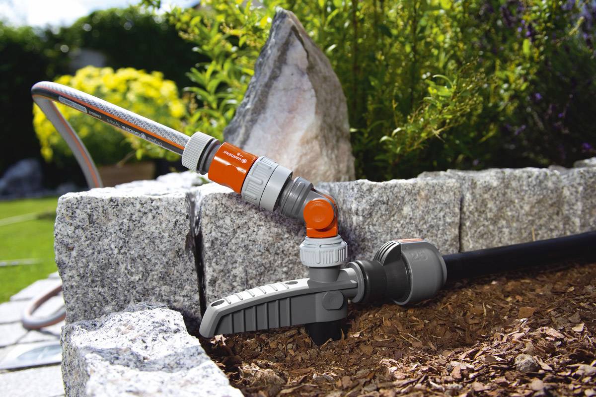 Un tuyau d'arrosage est raccordé à une pompe à eau installée dans un jardin, à côté de dalles de pierre et de paillis.