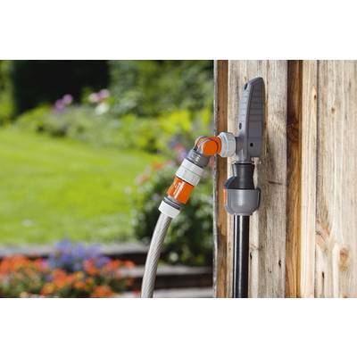 Un tuyau d'arrosage avec des raccords gris et orange est connecté à un robinet sur un mur en bois, avec un jardin en arrière-plan.