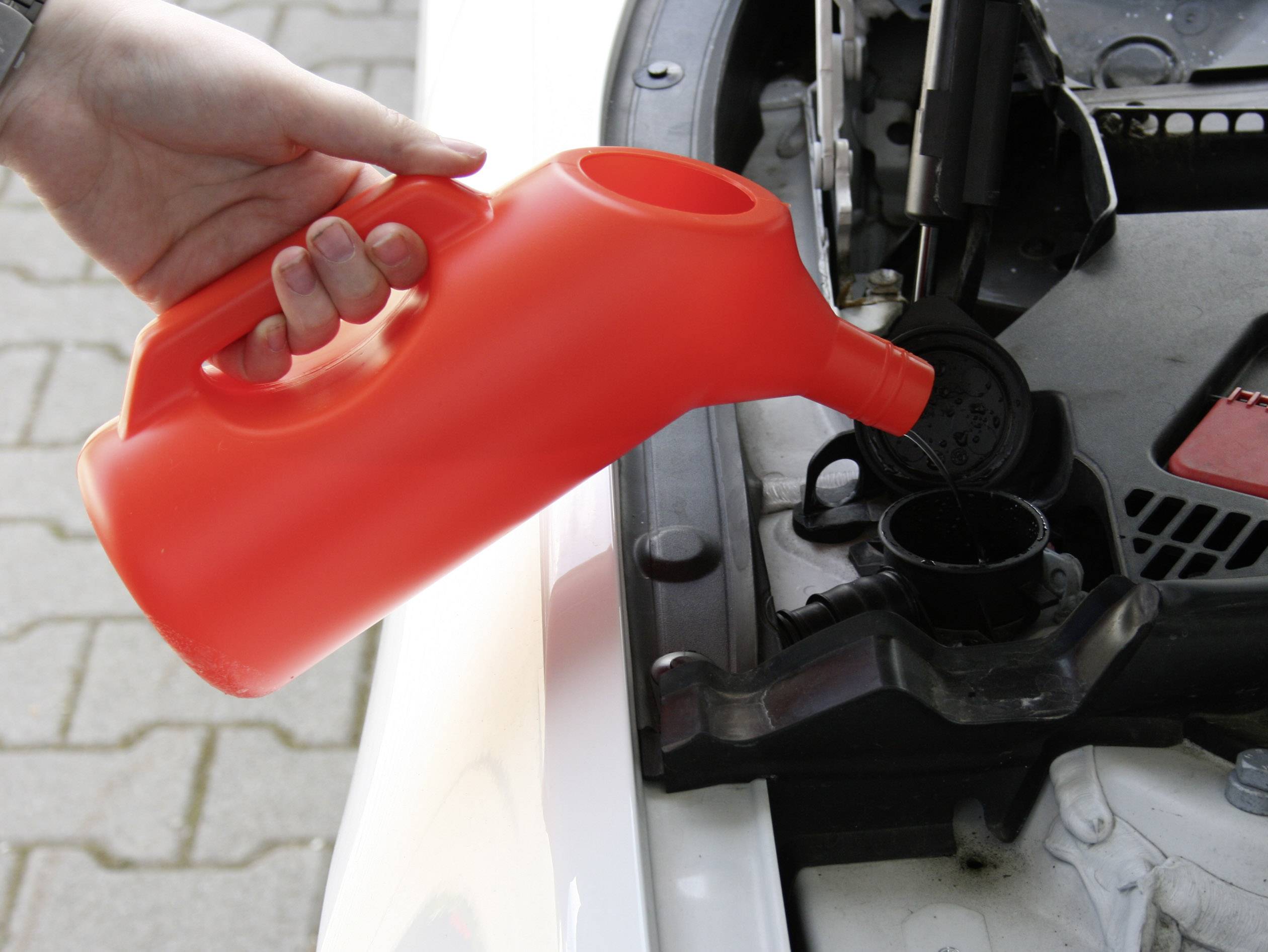 Une personne verse du liquide de refroidissement d'un bidon rouge dans le moteur d'une voiture. Le compartiment moteur est partiellement visible.