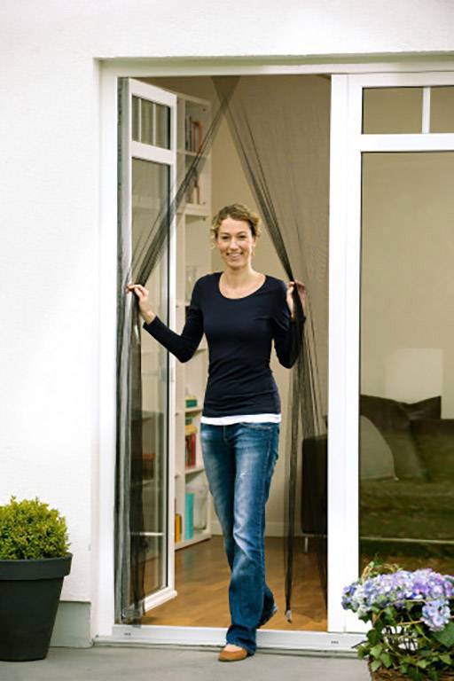 Une personne souriante se tient sur le balcon ouvert, écartant une moustiquaire. À l'extérieur, des plantes sont disposées près de la porte.