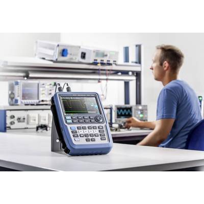 Un technicien est assis dans un laboratoire et travaille avec un oscilloscope posé sur une table. D'autres équipements électroniques sont visibles en arrière-plan.
