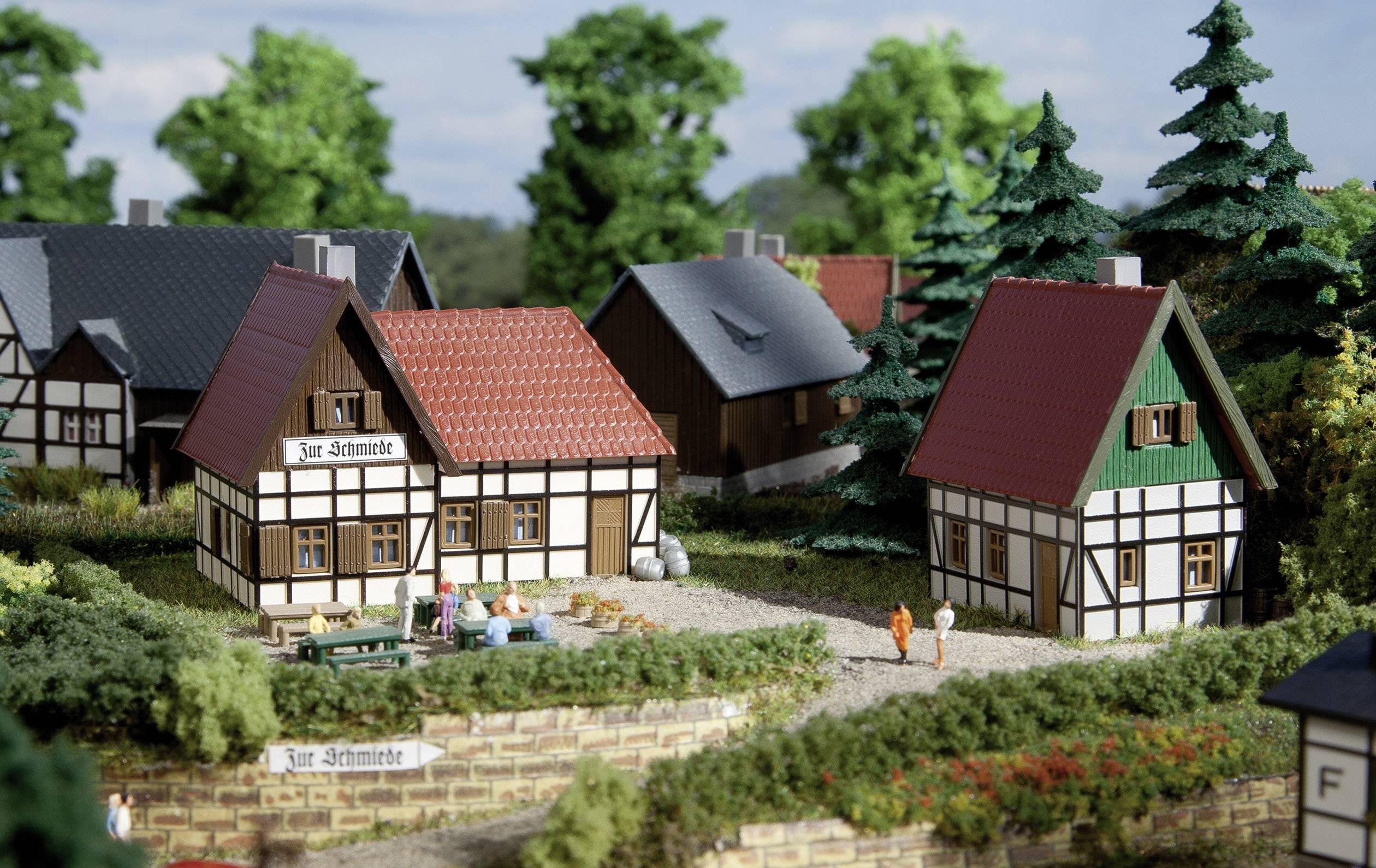 Petit village miniature avec des maisons à colombages, des jardins et des personnages sur un chemin. Maisons aux toits rouges et gris, arbres en arrière-plan.